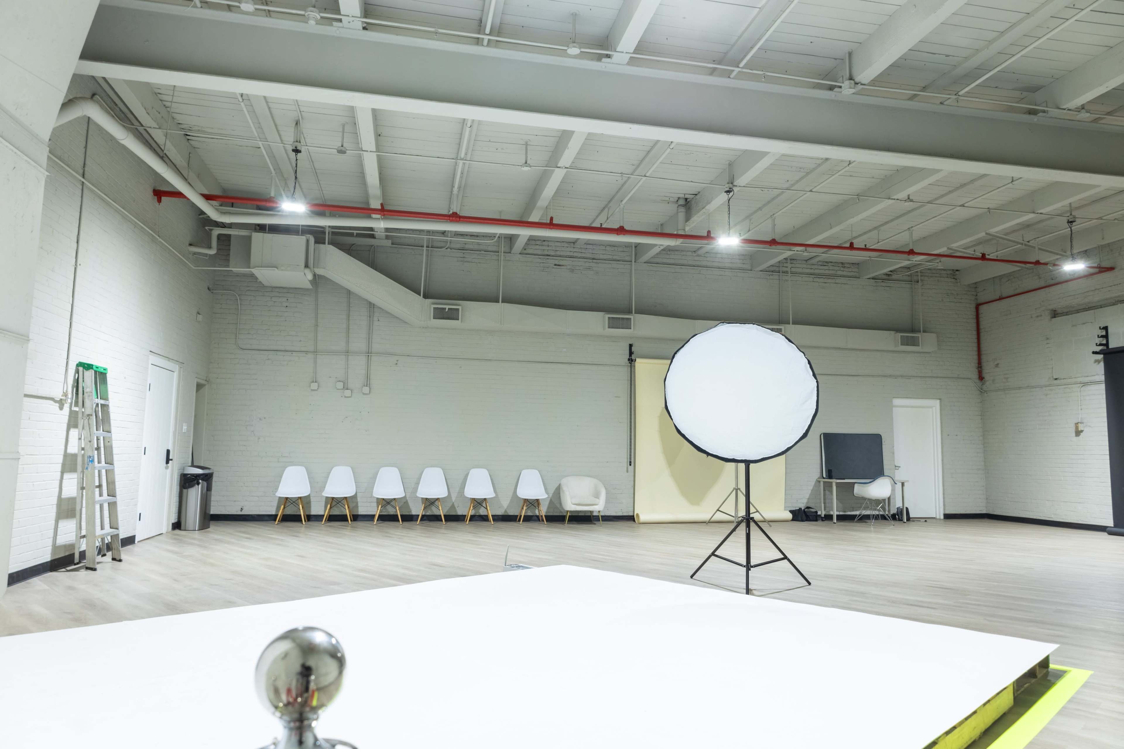The image shows a spacious, empty studio with a large white backdrop stand, several white chairs lined up against one wall, and a ladder in one corner.
