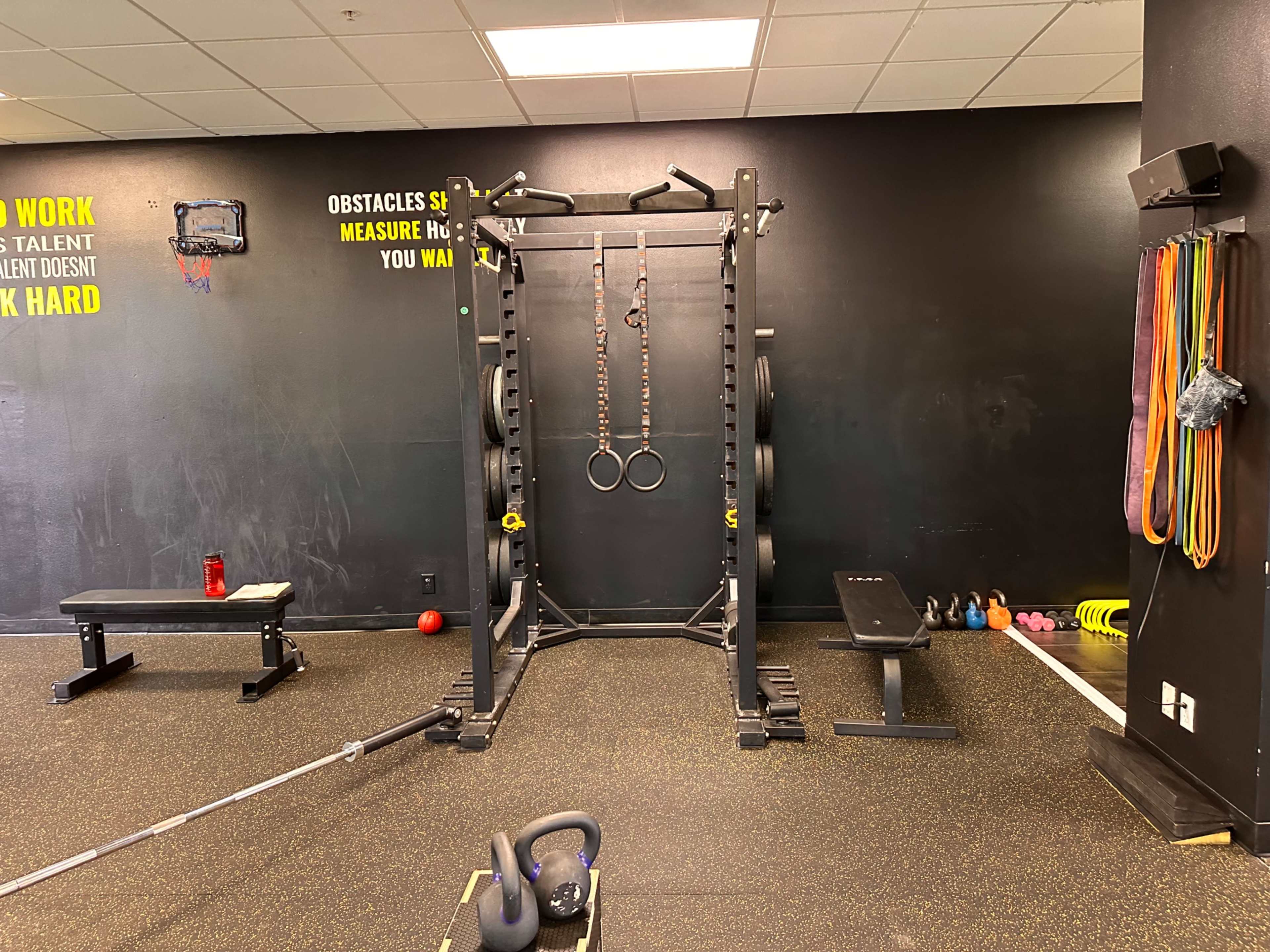 A gym with a squat rack featuring gym rings, a bench, and various weights and equipment against a black wall.