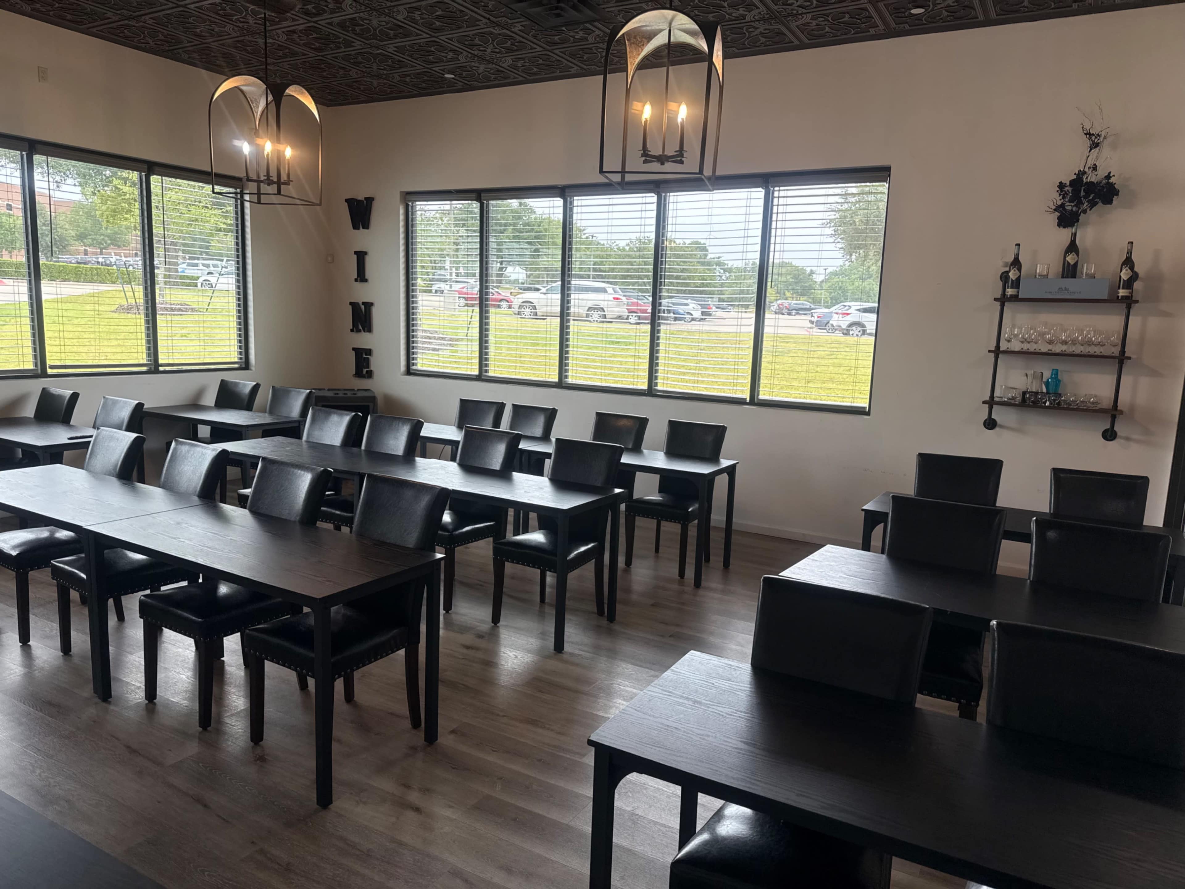 The image shows a spacious dining area with black tables and chairs, large windows providing natural light, and a wall shelf displaying decorative items.