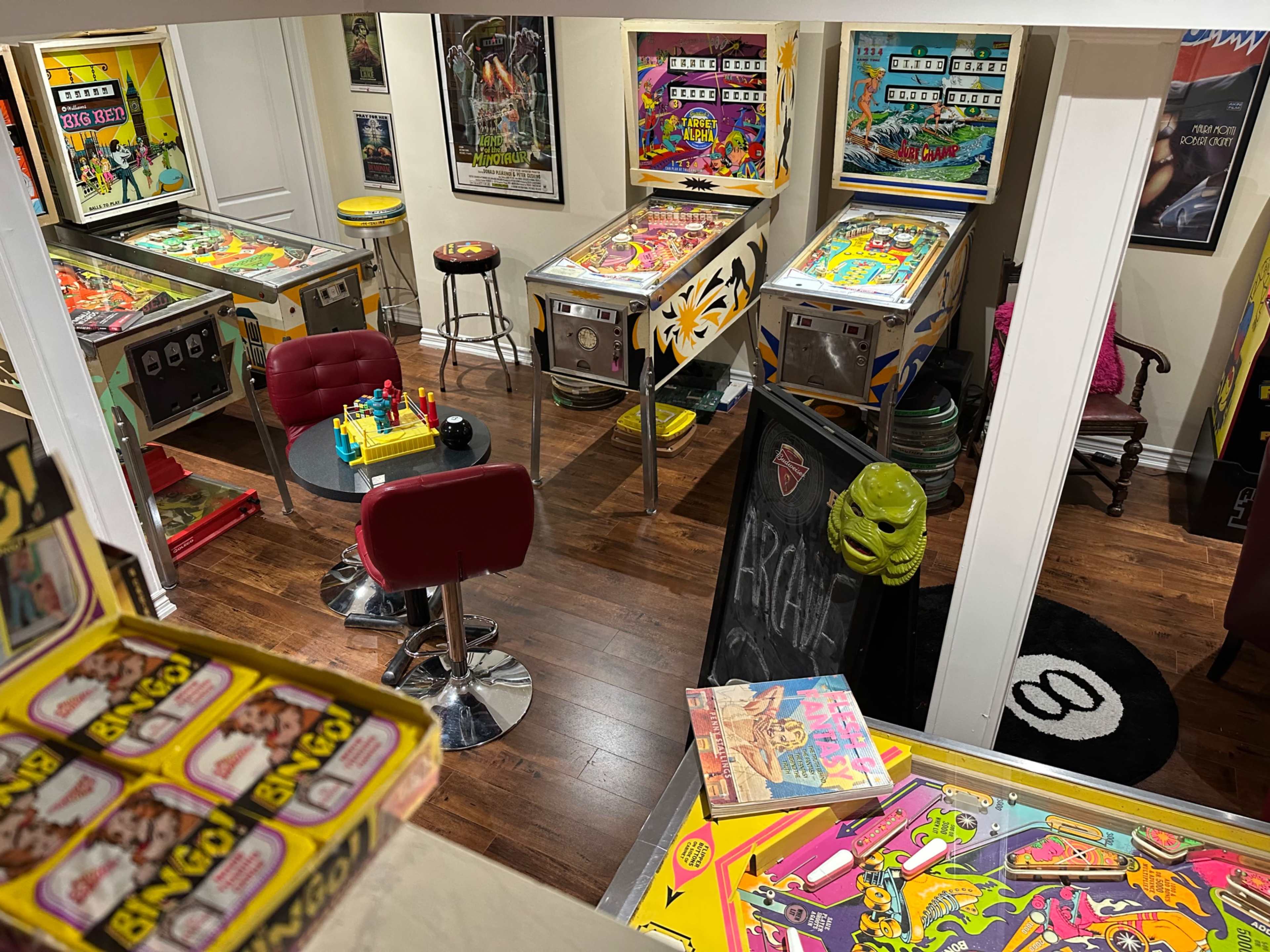 The image shows a room filled with vintage pinball machines, game tables, and seating areas, showcasing a colorful and nostalgic arcade atmosphere.