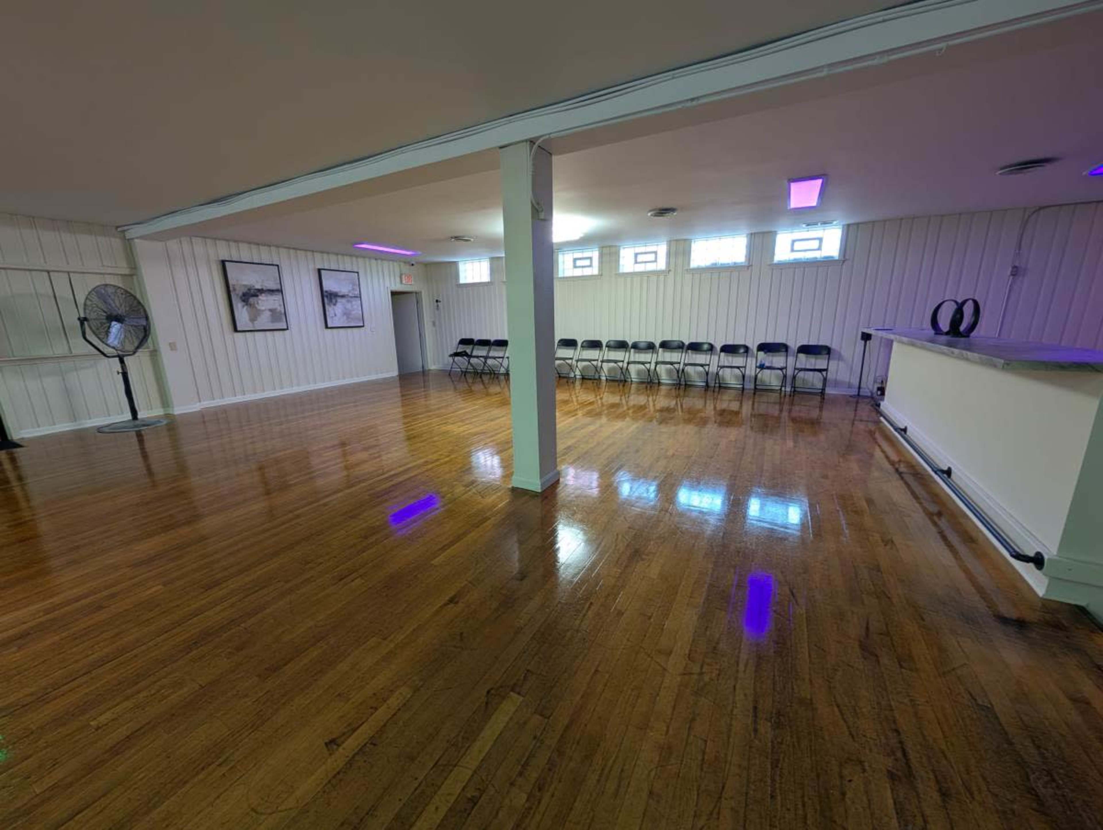The image shows a spacious, empty room with polished wooden floors, a row of black chairs along one side, and large windows near the ceiling.