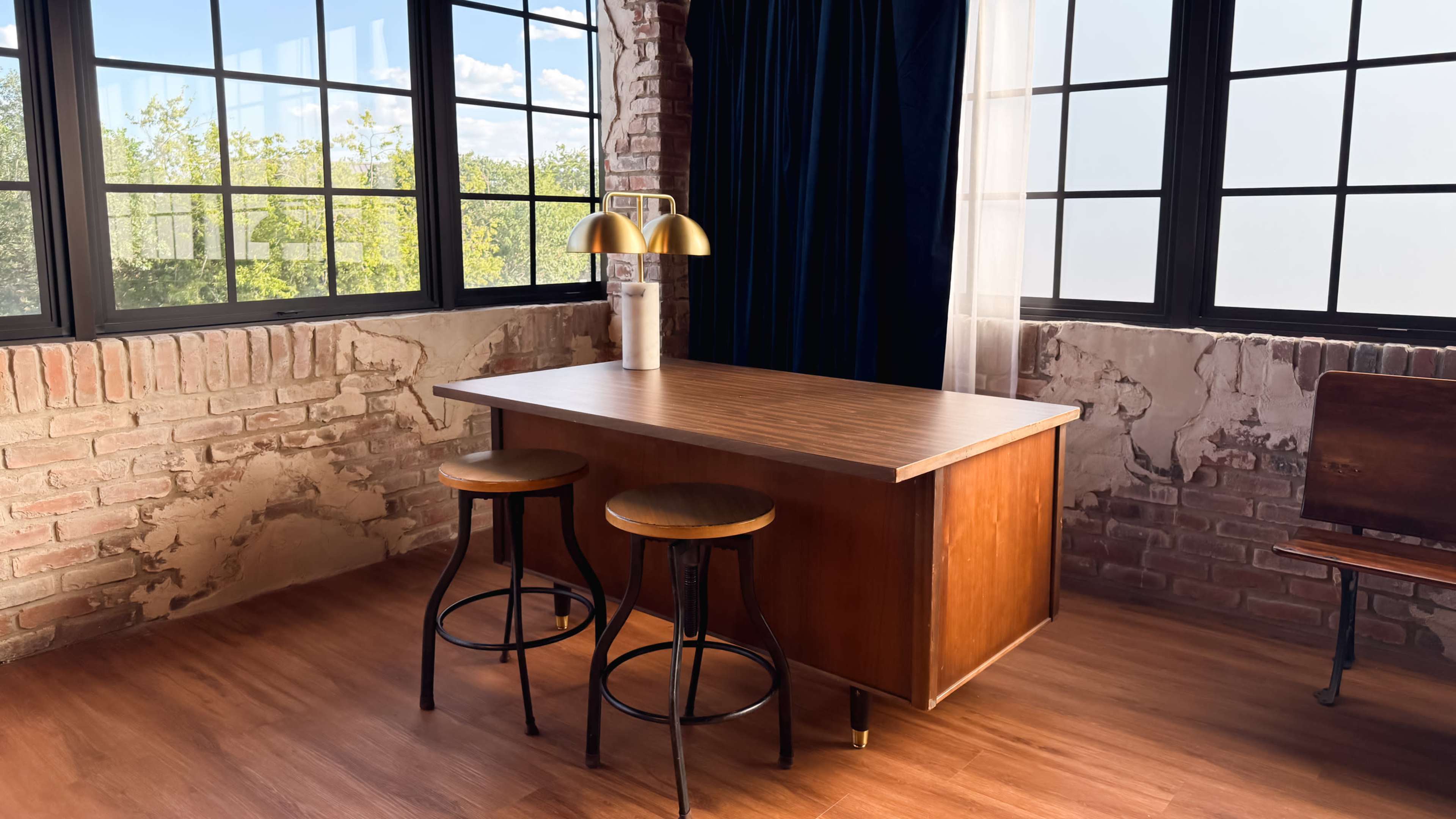 A wooden table with two black stools is placed against a wall with exposed brick, near large windows that overlook a green landscape.