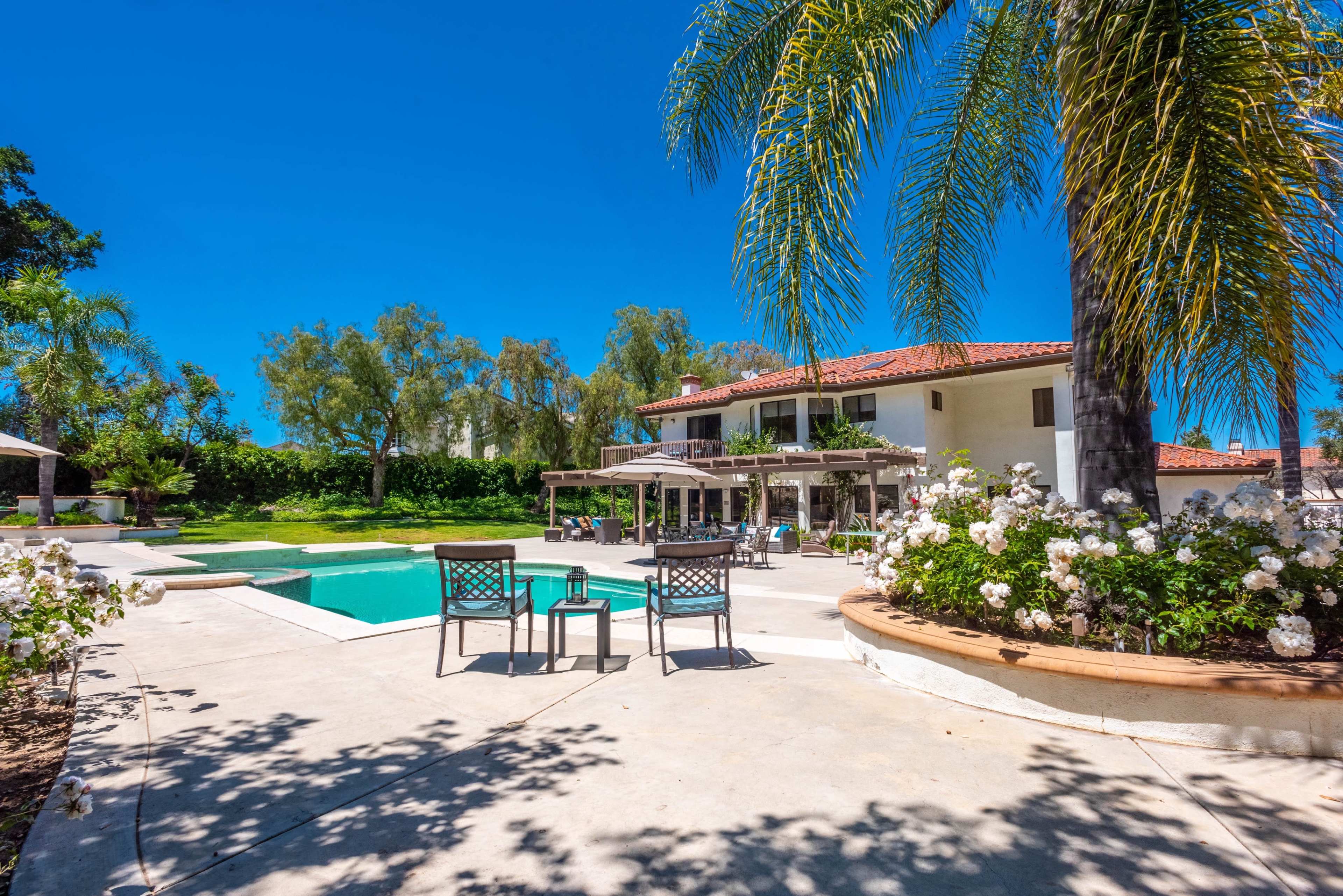 A spacious backyard features a swimming pool surrounded by lounge chairs and shaded areas near a large house with a red-tiled roof and landscaped gardens.