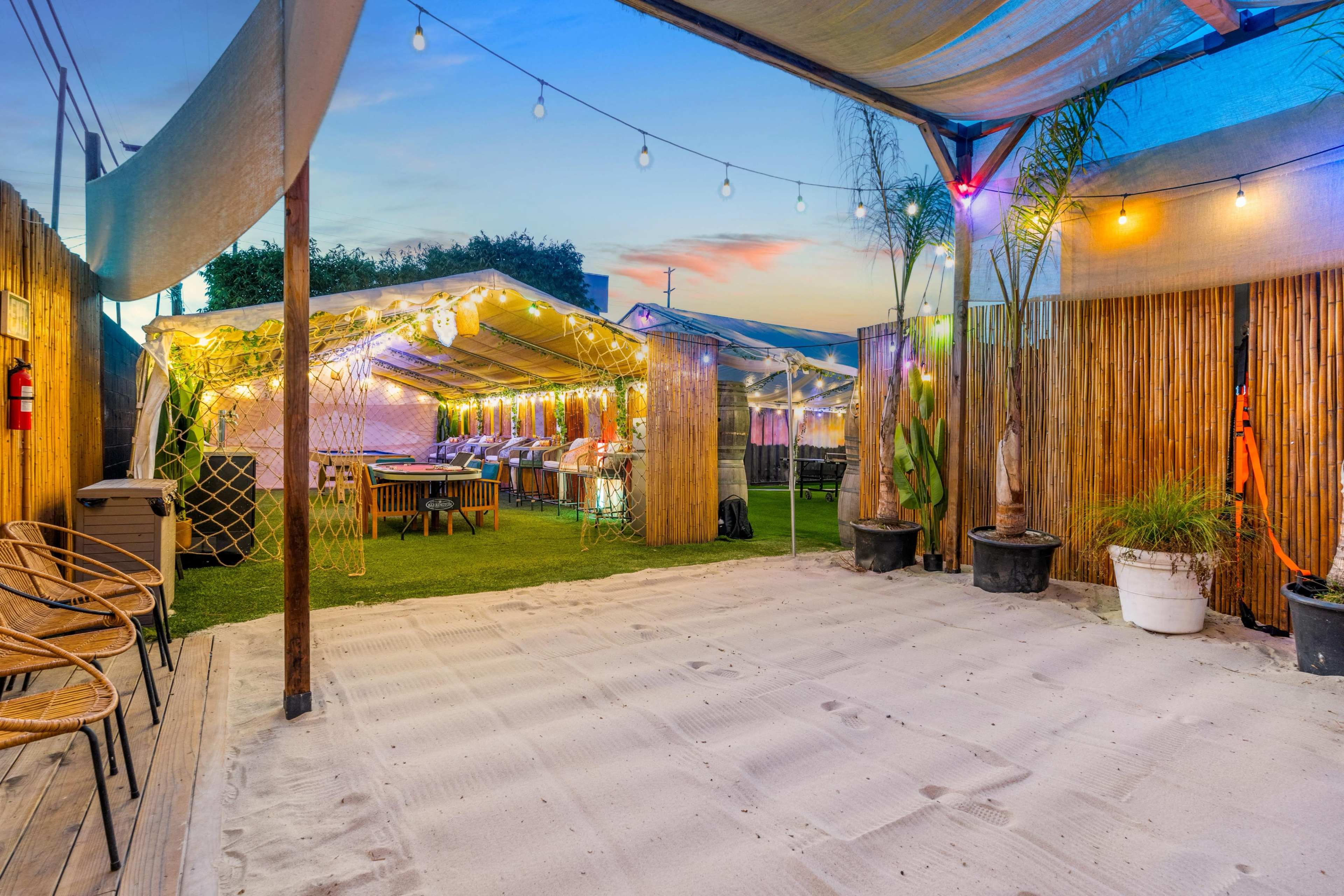 The image shows an outdoor event space with tables and string lights set up under tents amidst decorative plants.