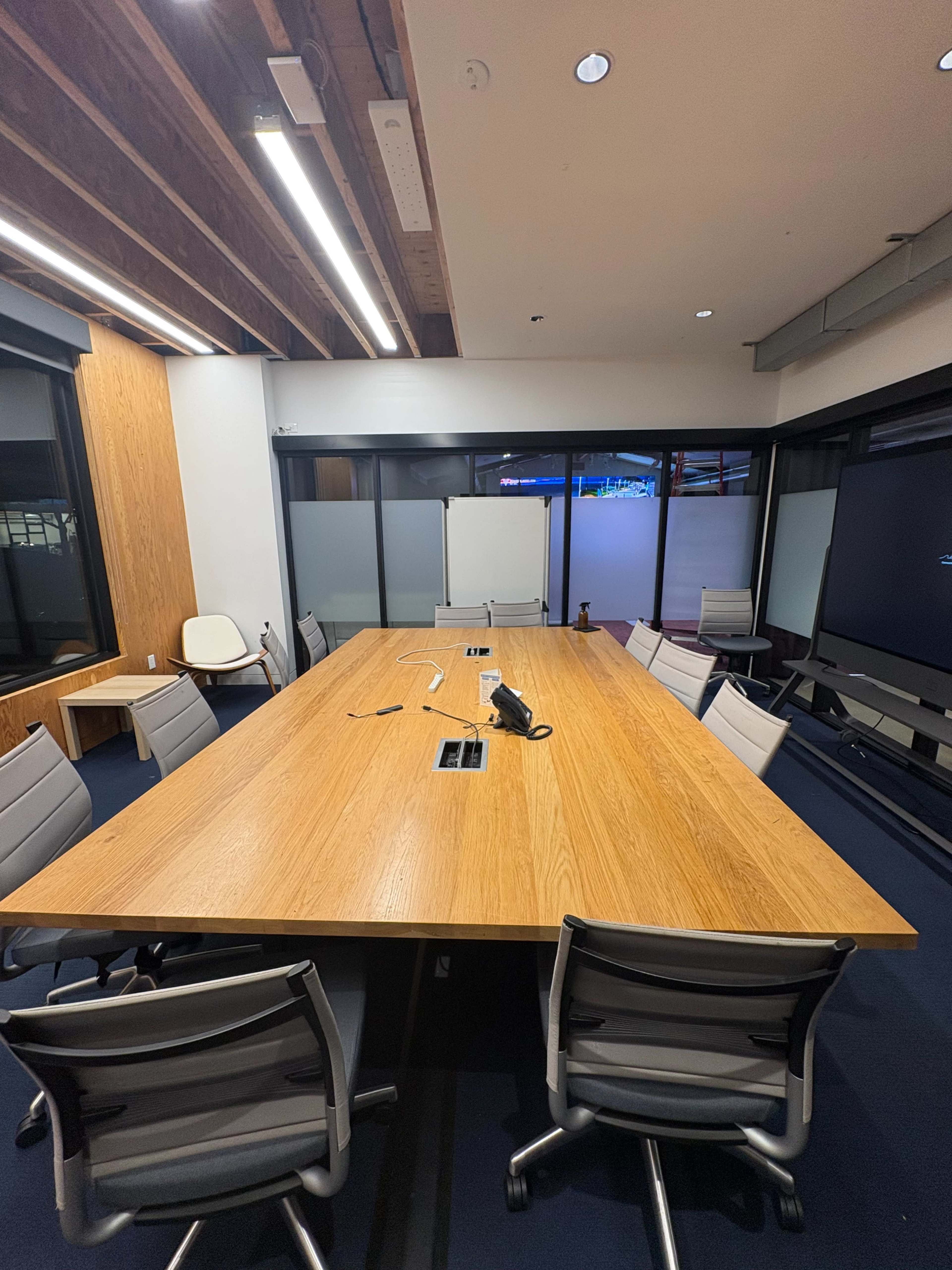 A large wooden conference table is set in a modern meeting room with several ergonomic chairs and a projector screen visible in the background.