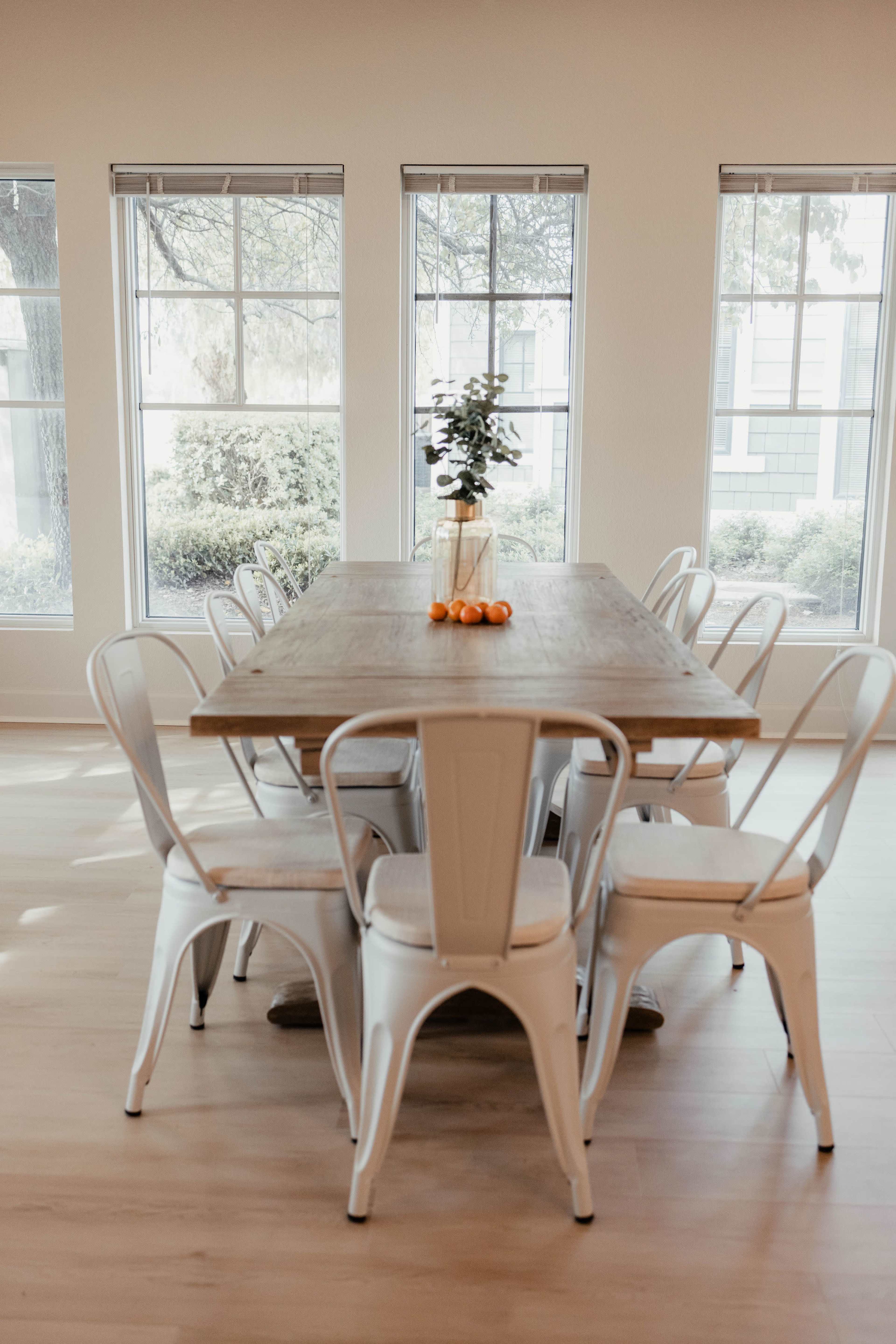A wooden dining table surrounded by white chairs is set in a well-lit room with large windows.