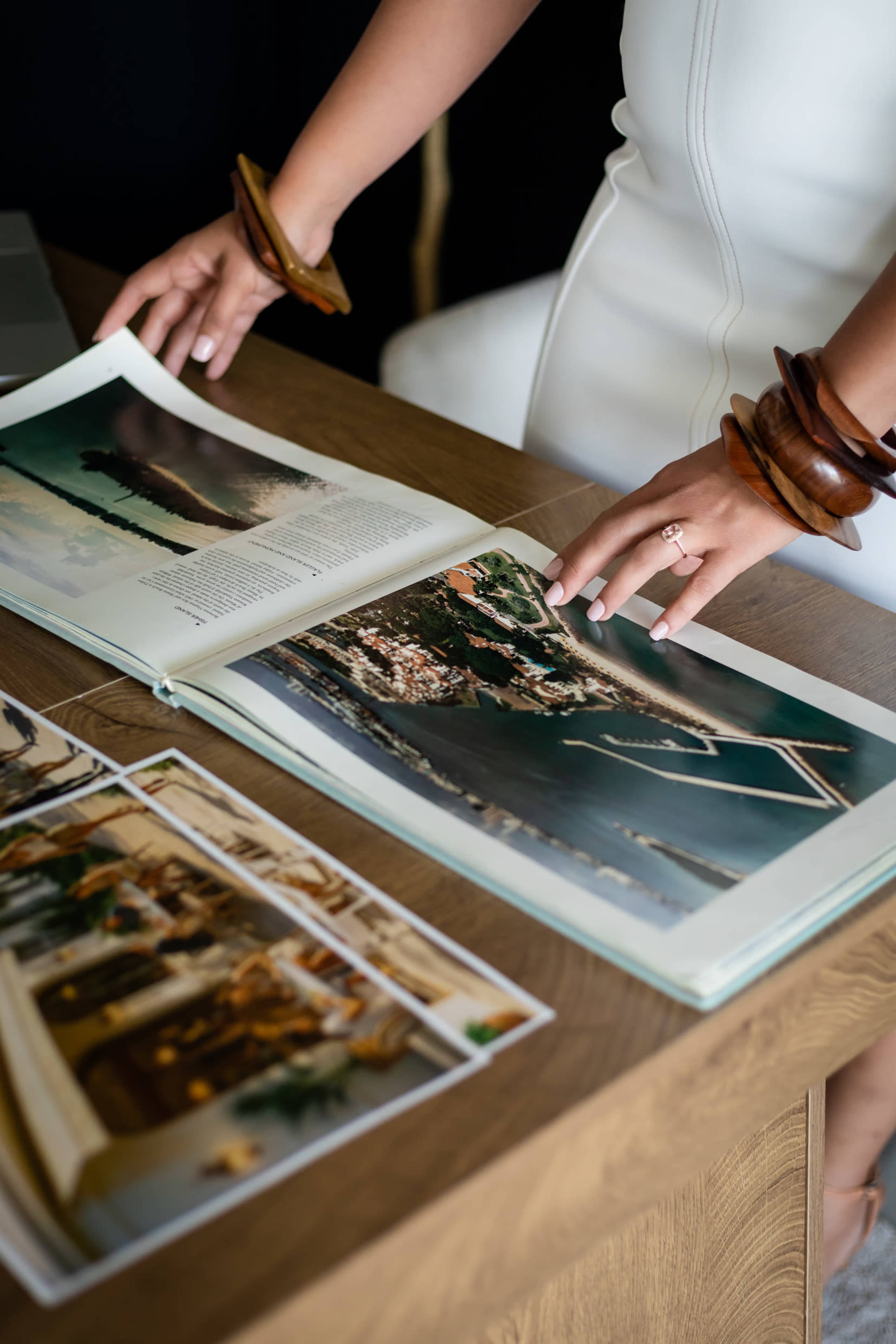 A person with wooden bangles examines an open book on a wooden table that features photographs of aerial views and landscapes.