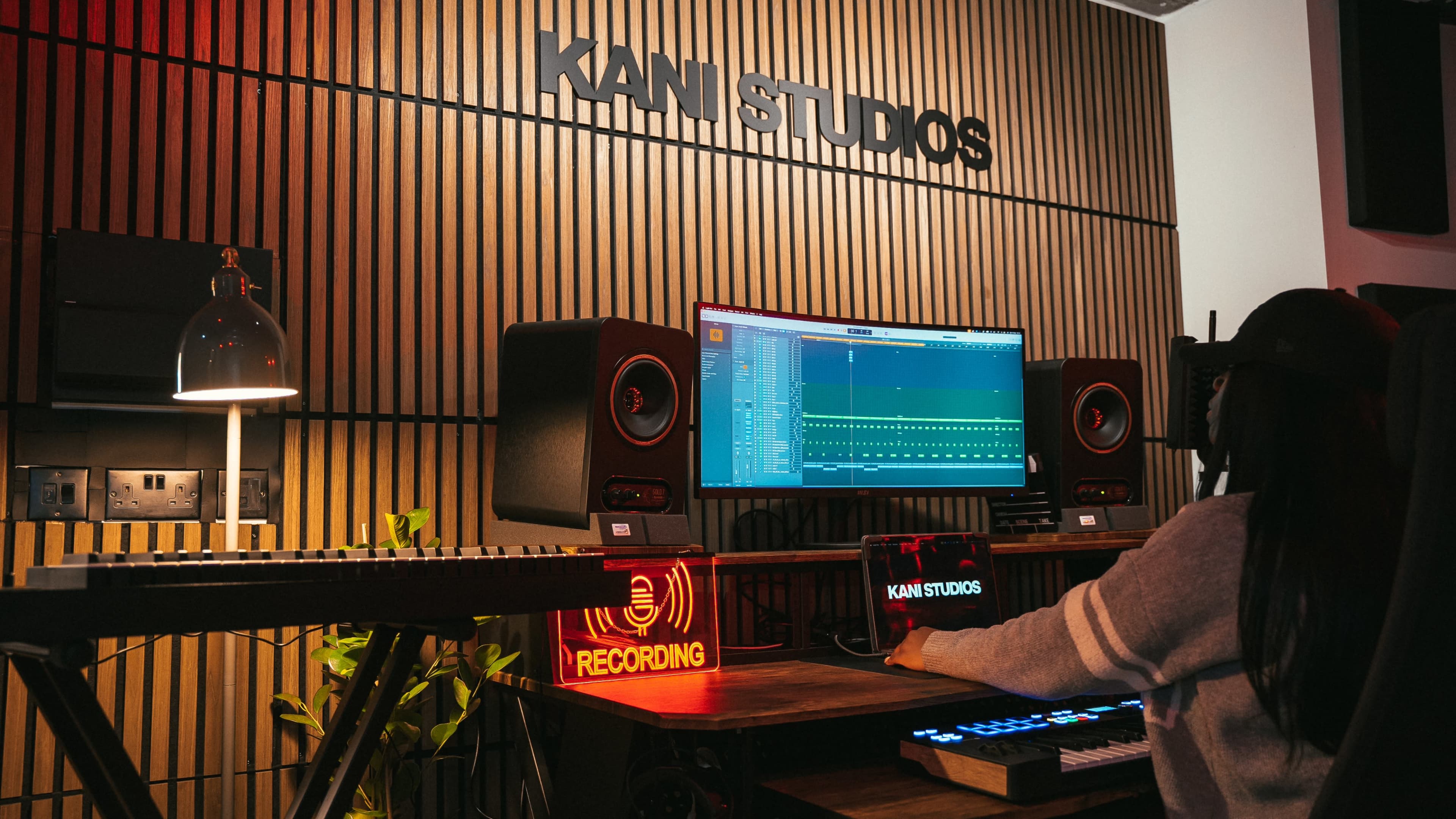 A musician works at a well-equipped recording studio, featuring a computer monitor displaying music software and various sound equipment, with "KANI STUDIOS" visible on the wall.
