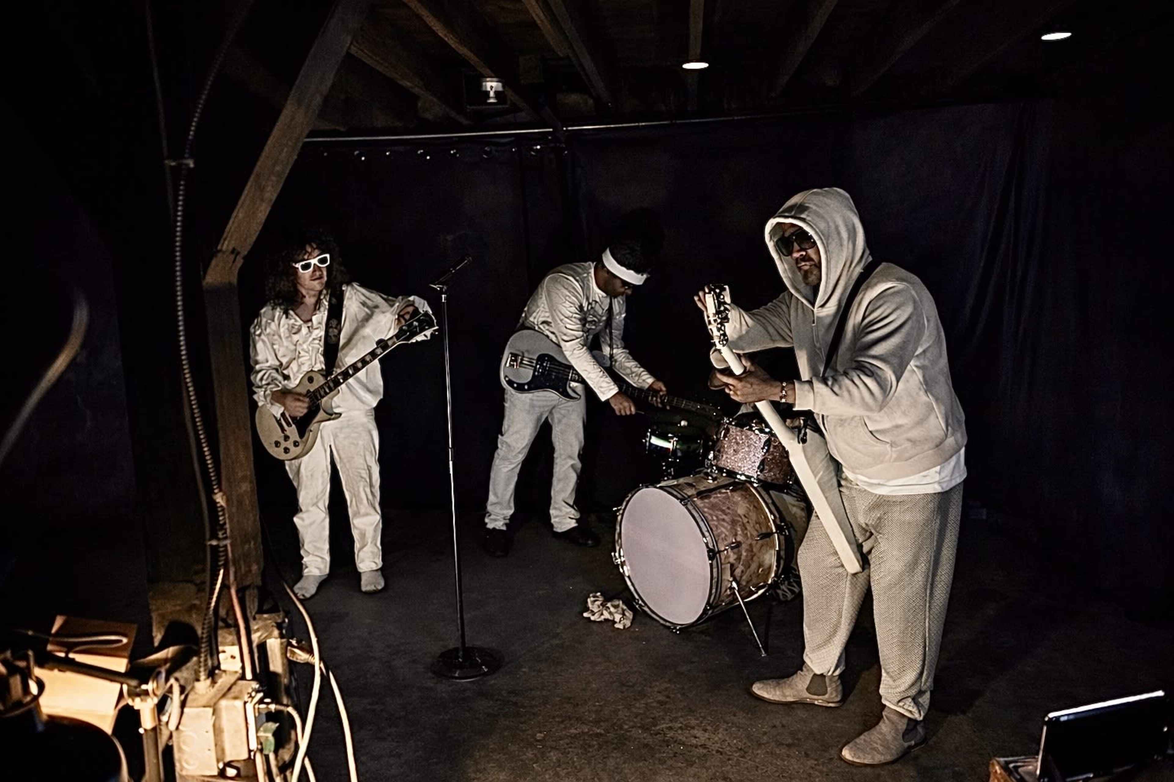 A band performs in a dimly lit basement, with one member playing guitar, another on drums, and the third singing into a microphone.
