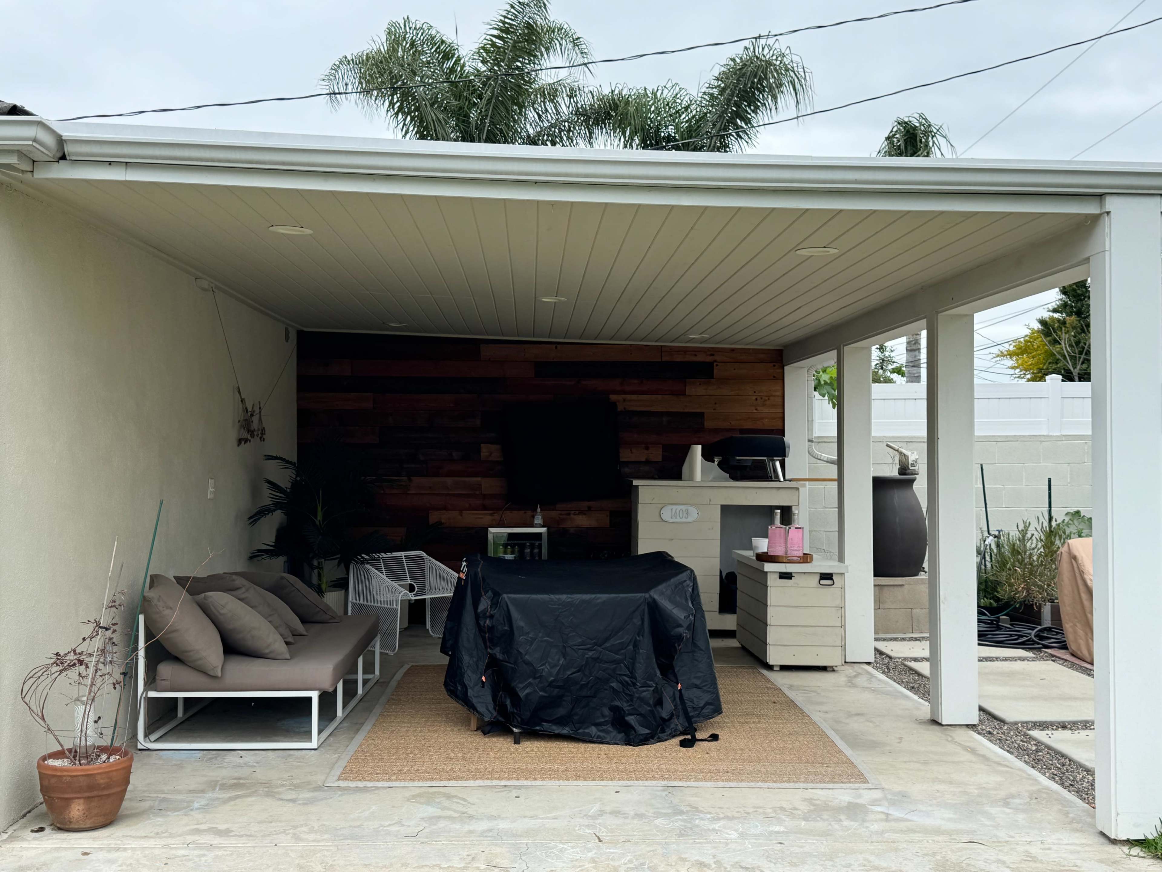 A covered outdoor patio with a seating area, a TV mounted on the wall, and a grill covered with a black tarp.