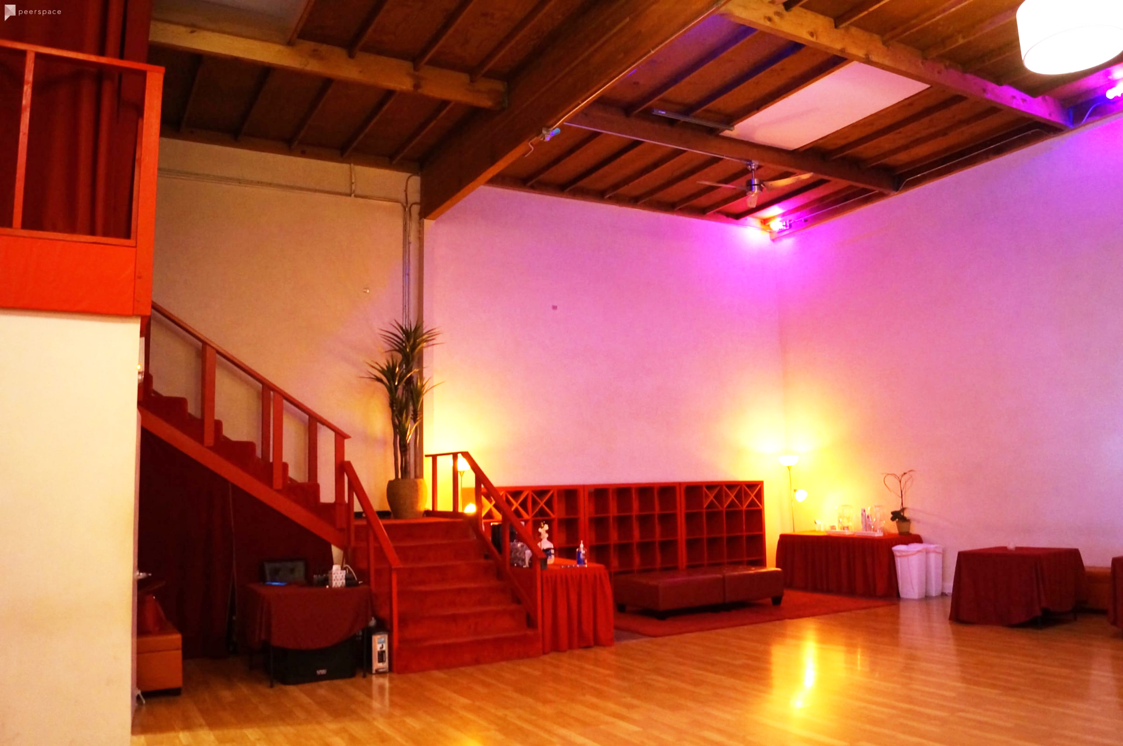 A spacious room with wooden floors, a staircase leading to an upper level, and red curtains and furniture, illuminated by soft colored lighting.