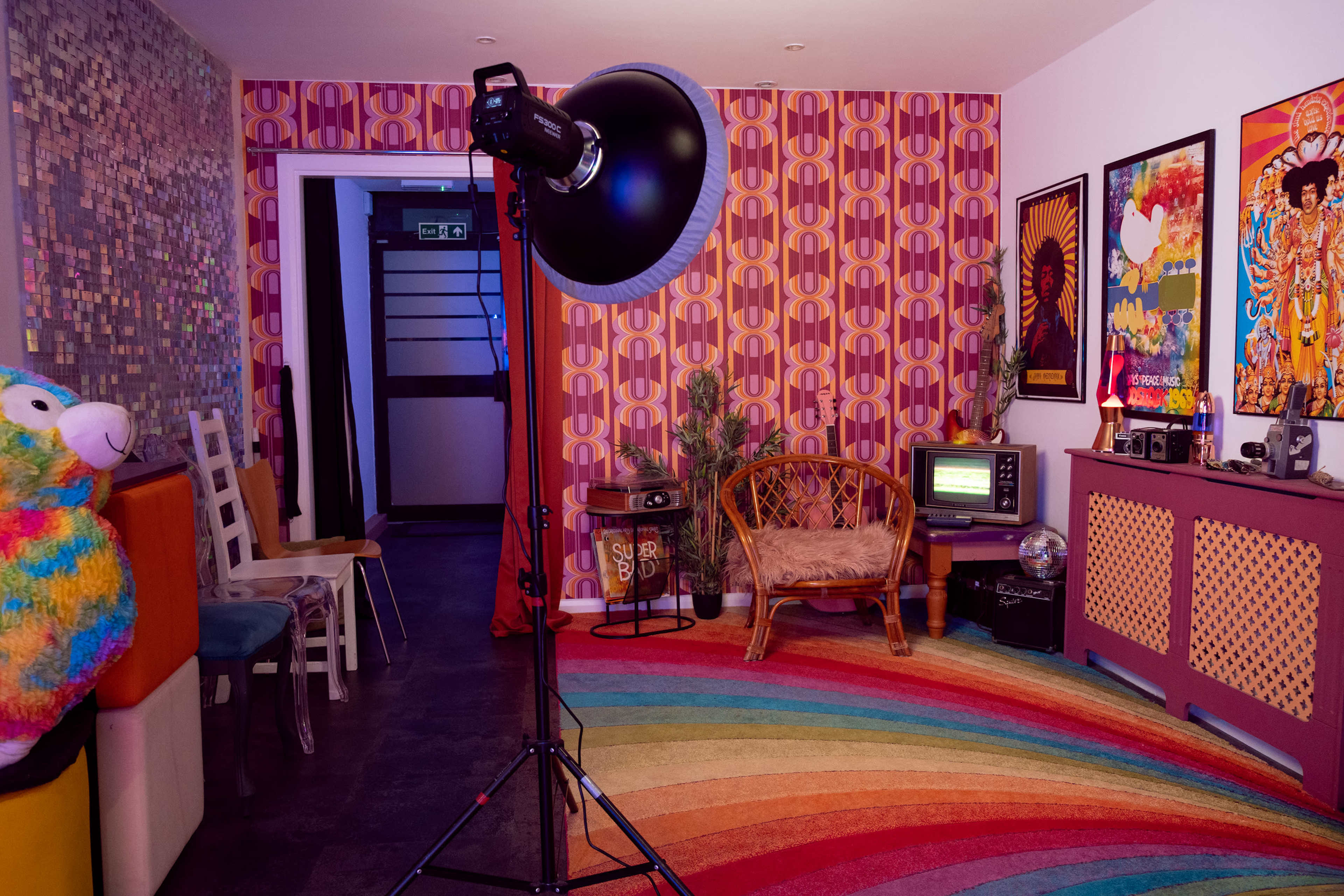 The image shows a colorful room with a large light stand, a vintage television, a peacock chair, and a vibrant, circular rug.