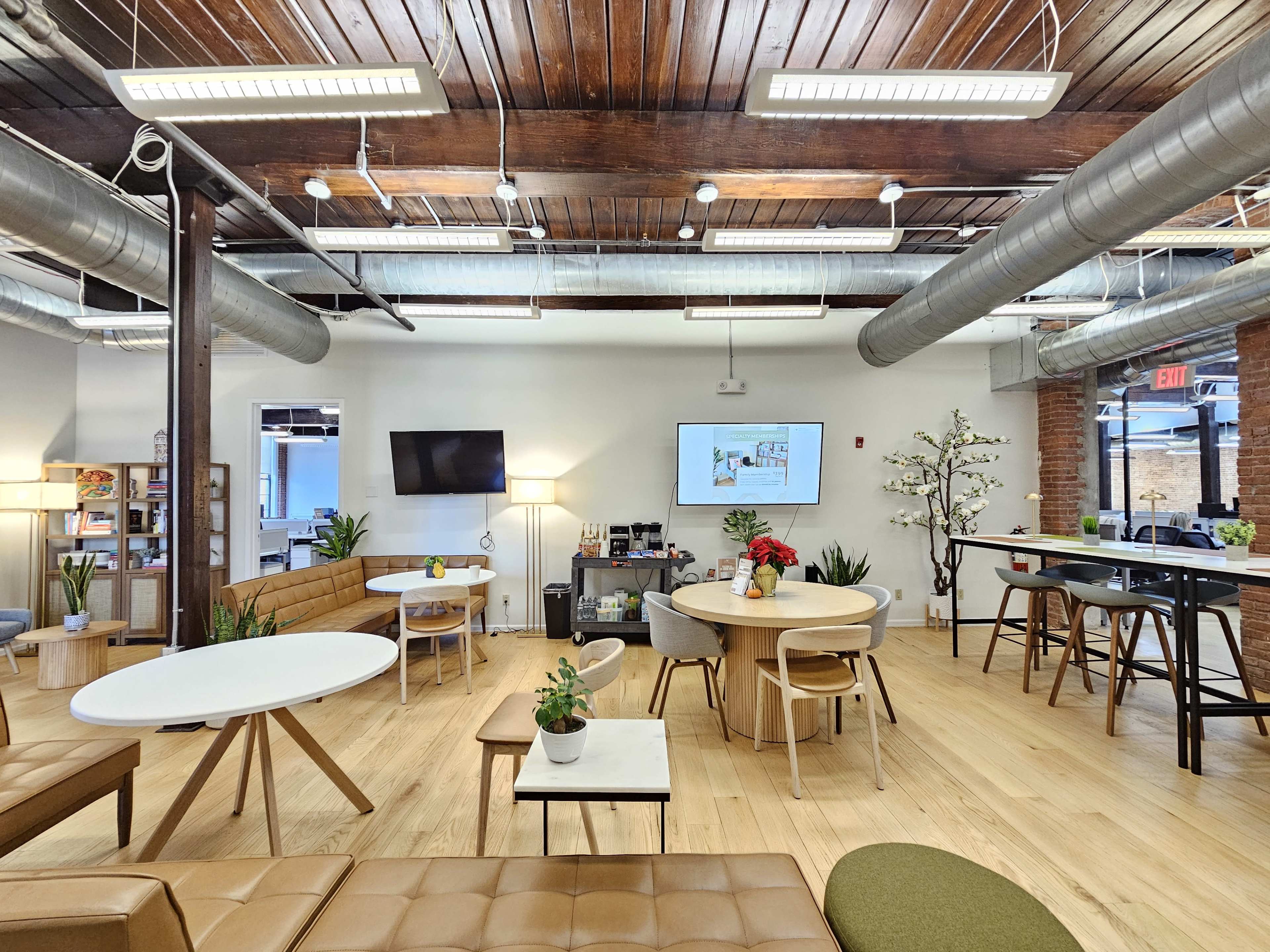 The image shows a modern office workspace with wooden floors, various tables and seating arrangements, and wall-mounted screens.