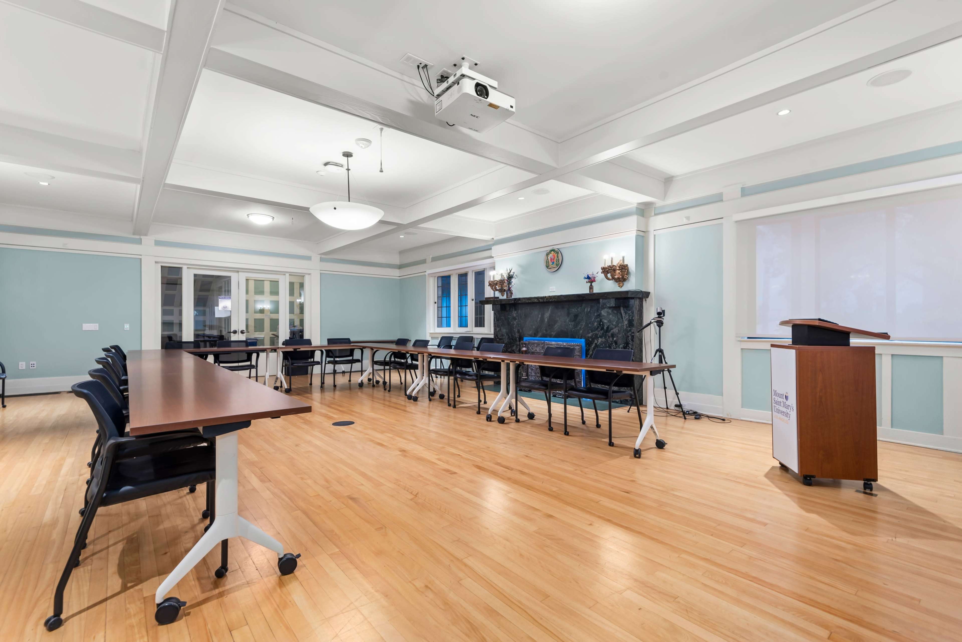 The image shows a spacious conference room with tables arranged in a U-shape, chairs around them, a podium, and a projector mounted on the ceiling.