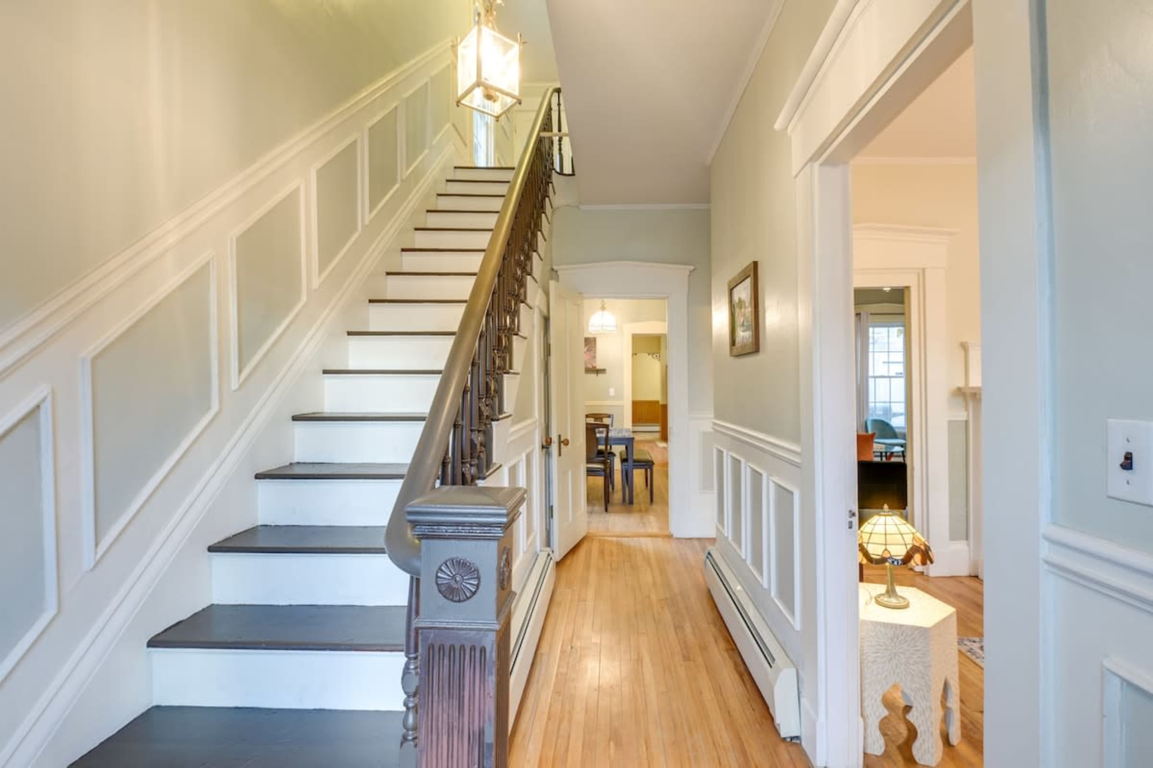 A well-lit hallway with a staircase, decorative wainscoting, and a view into a dining area beyond.
