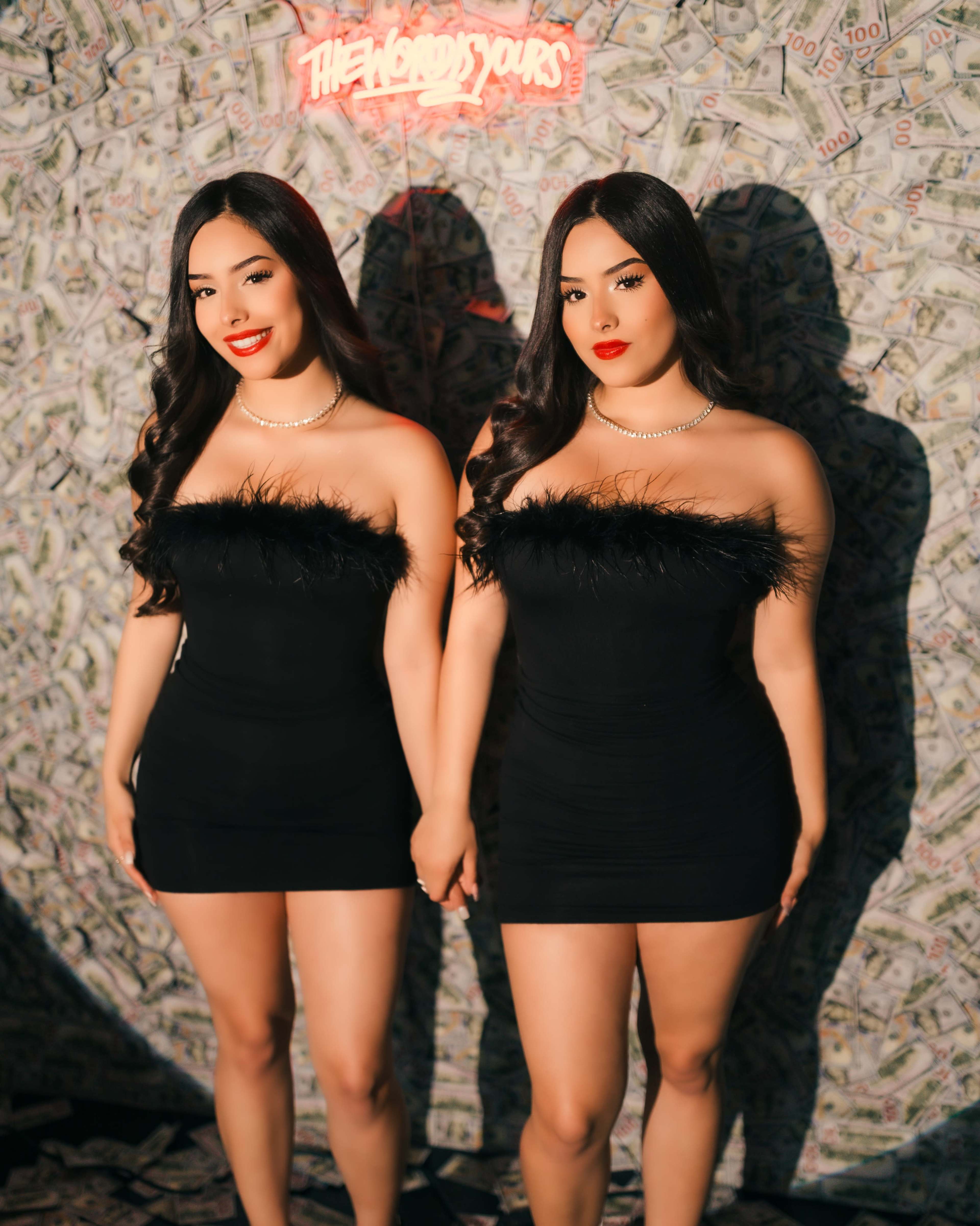Two women wearing identical black dresses with feather details pose together against a backdrop of money.