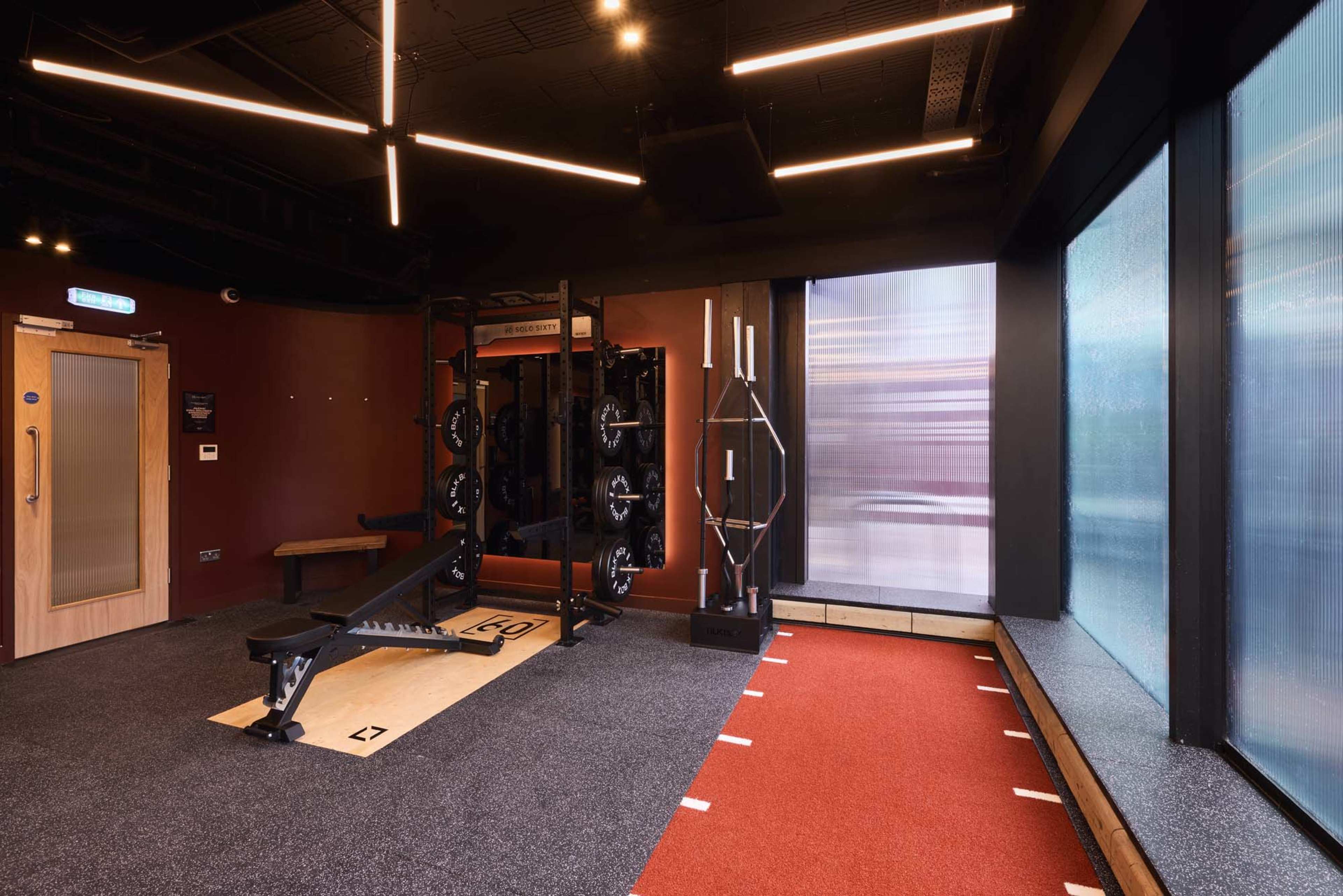 The image shows a modern gym interior featuring a weightlifting area with a bench, squat rack, and adjacent red turf for functional training.