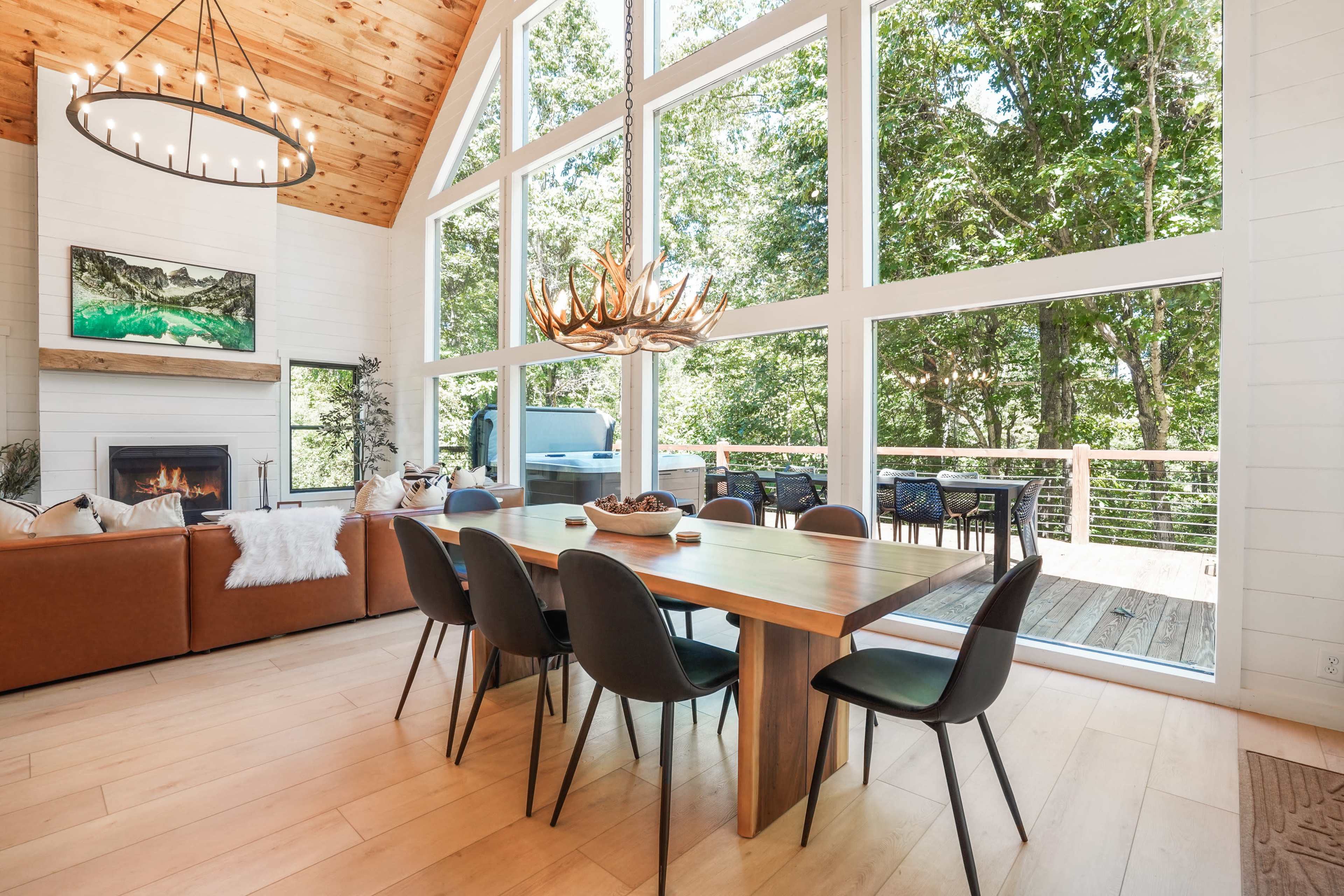 A bright dining area features a large wooden table surrounded by black chairs, with large windows overlooking a forested outdoor deck.