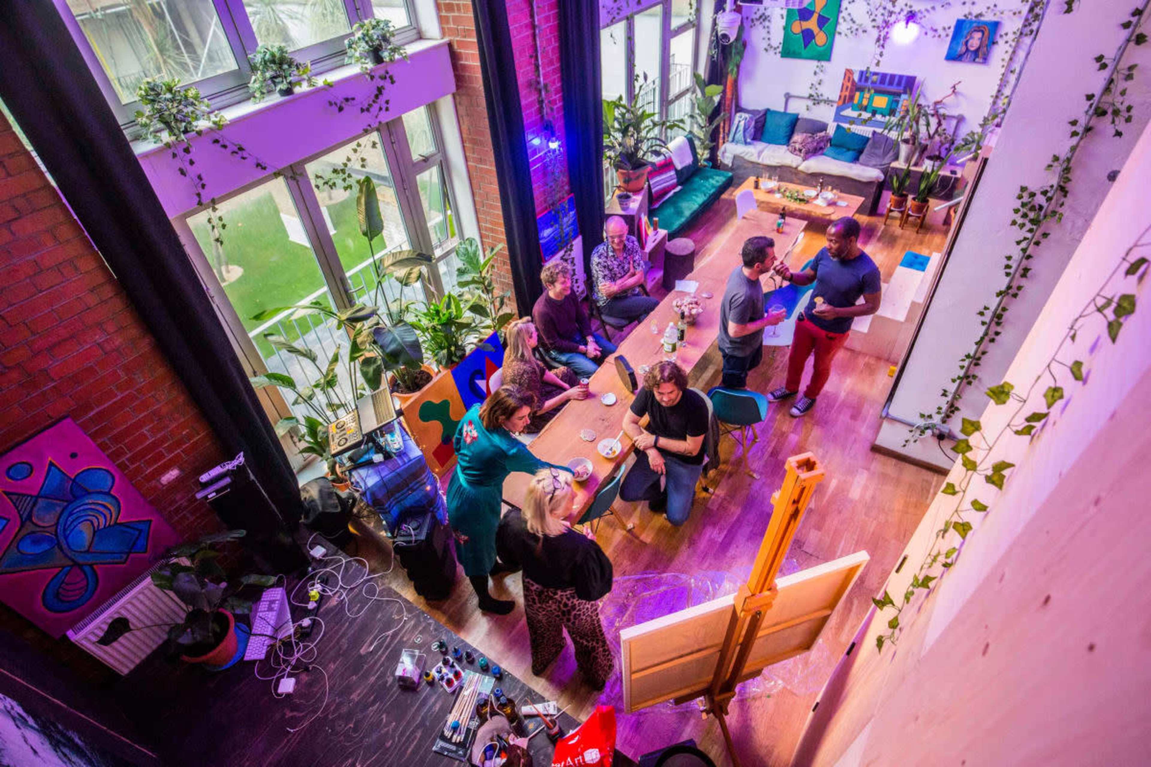 A group of people gather around a table in a colorful, plant-filled studio space while an artist works on a canvas nearby.