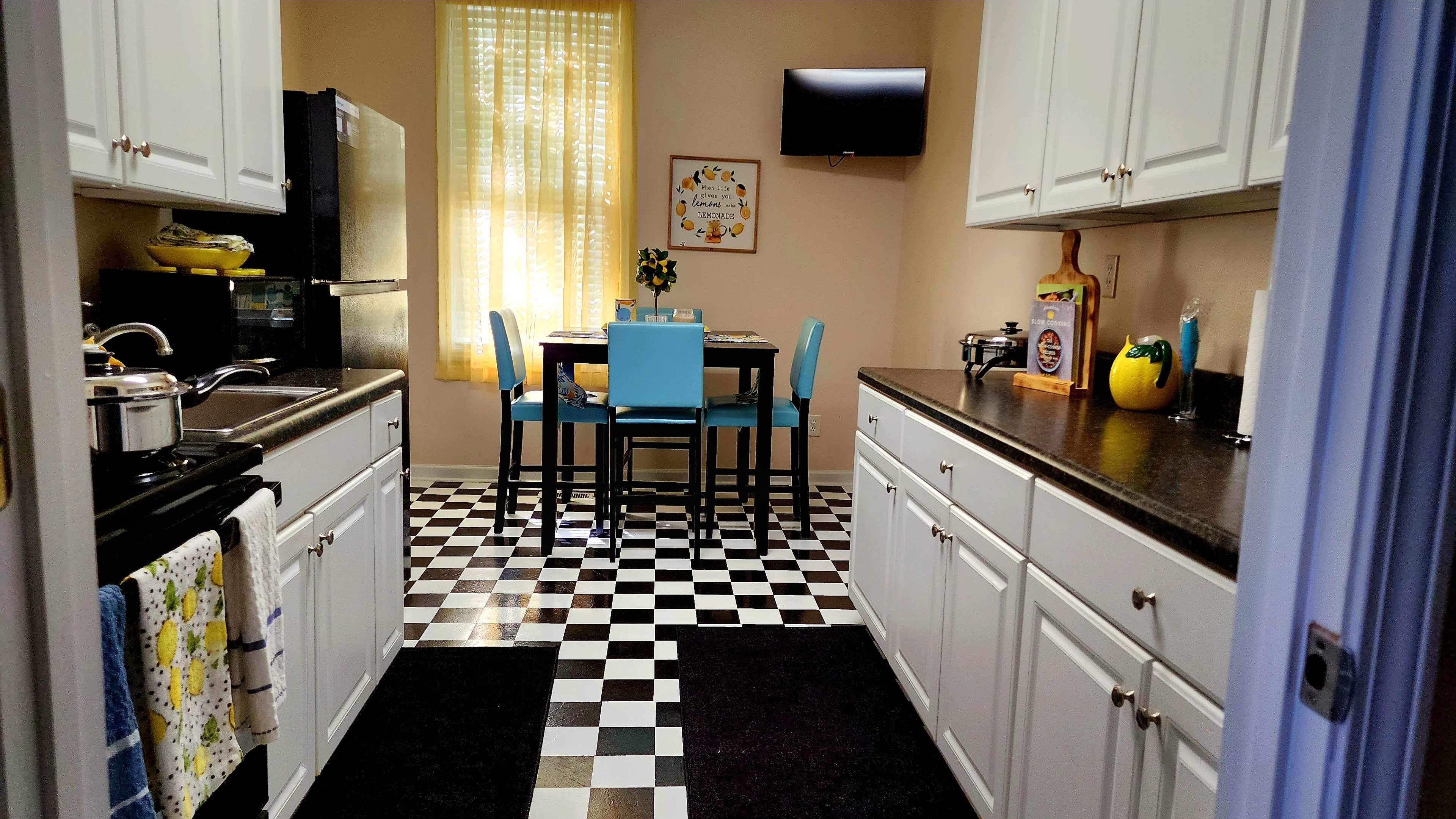A tidy kitchen features white cabinets, black and white checkerboard flooring, a dining table with blue chairs, and a black refrigerator.