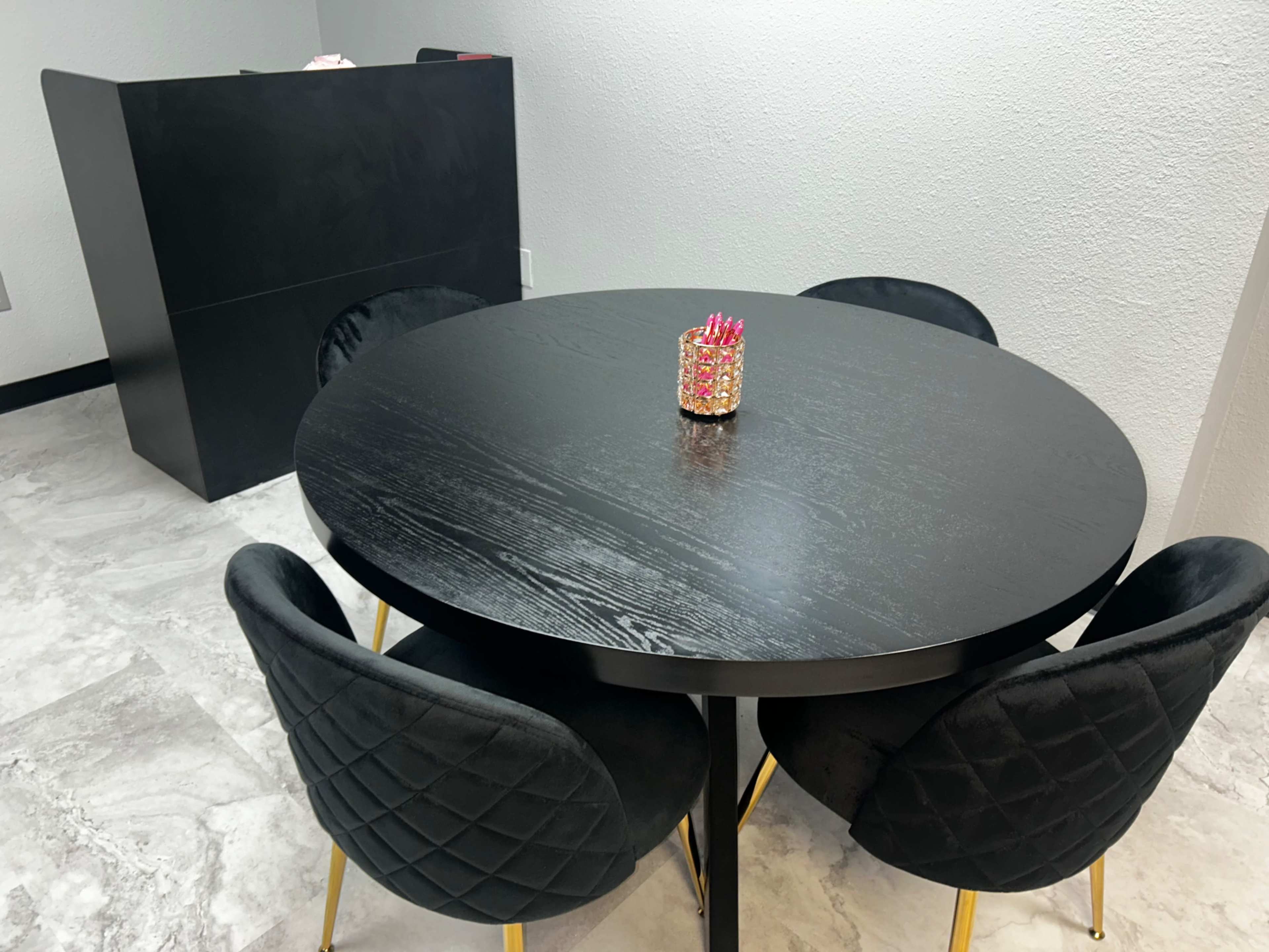A round black dining table with four black chairs and a decorative piece in the center is positioned in a room with light-colored flooring and a black reception desk in the background.