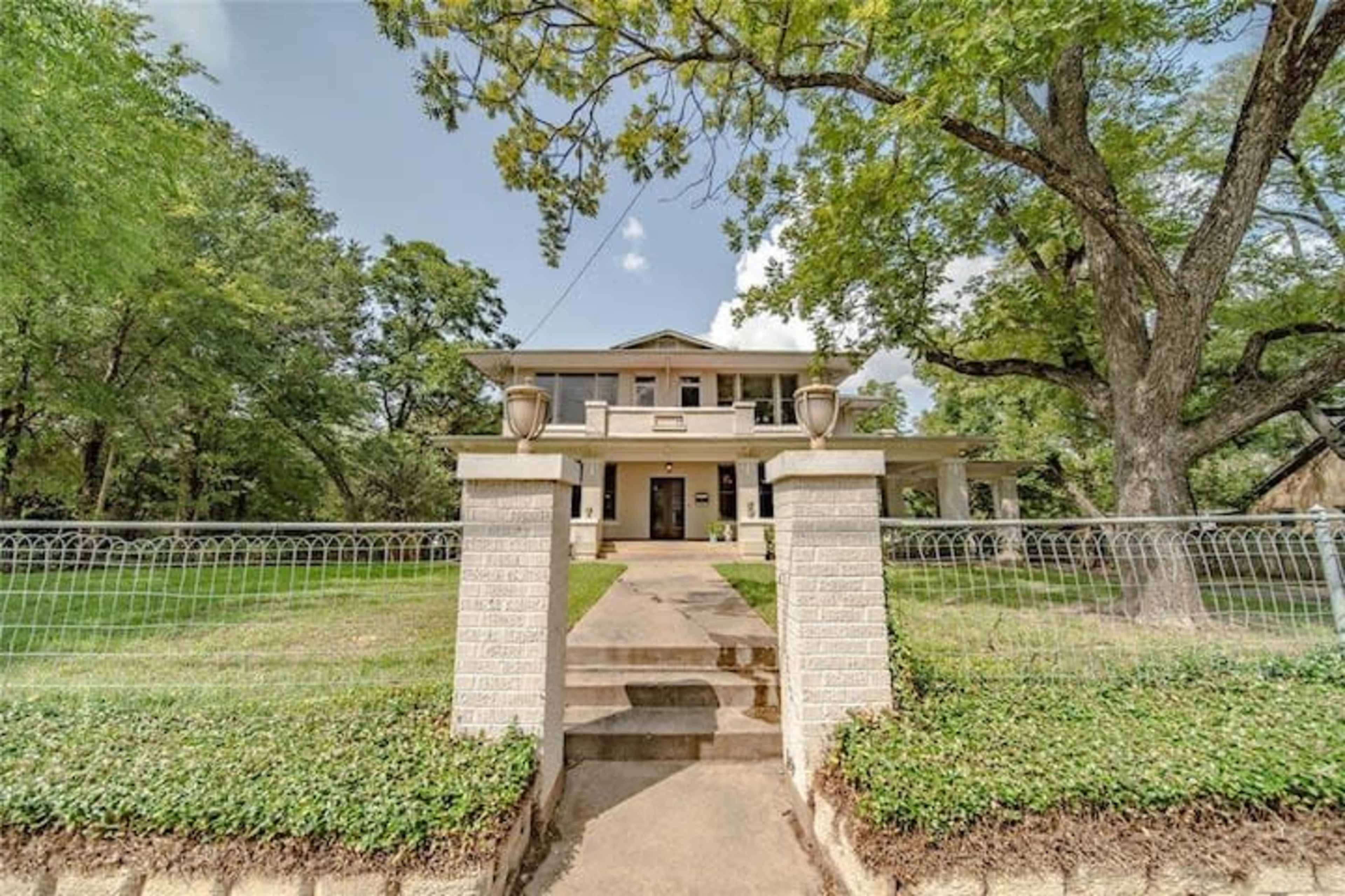 A two-story house with a large front porch is surrounded by trees and a decorative fence, featuring a pathway leading up to the entrance.