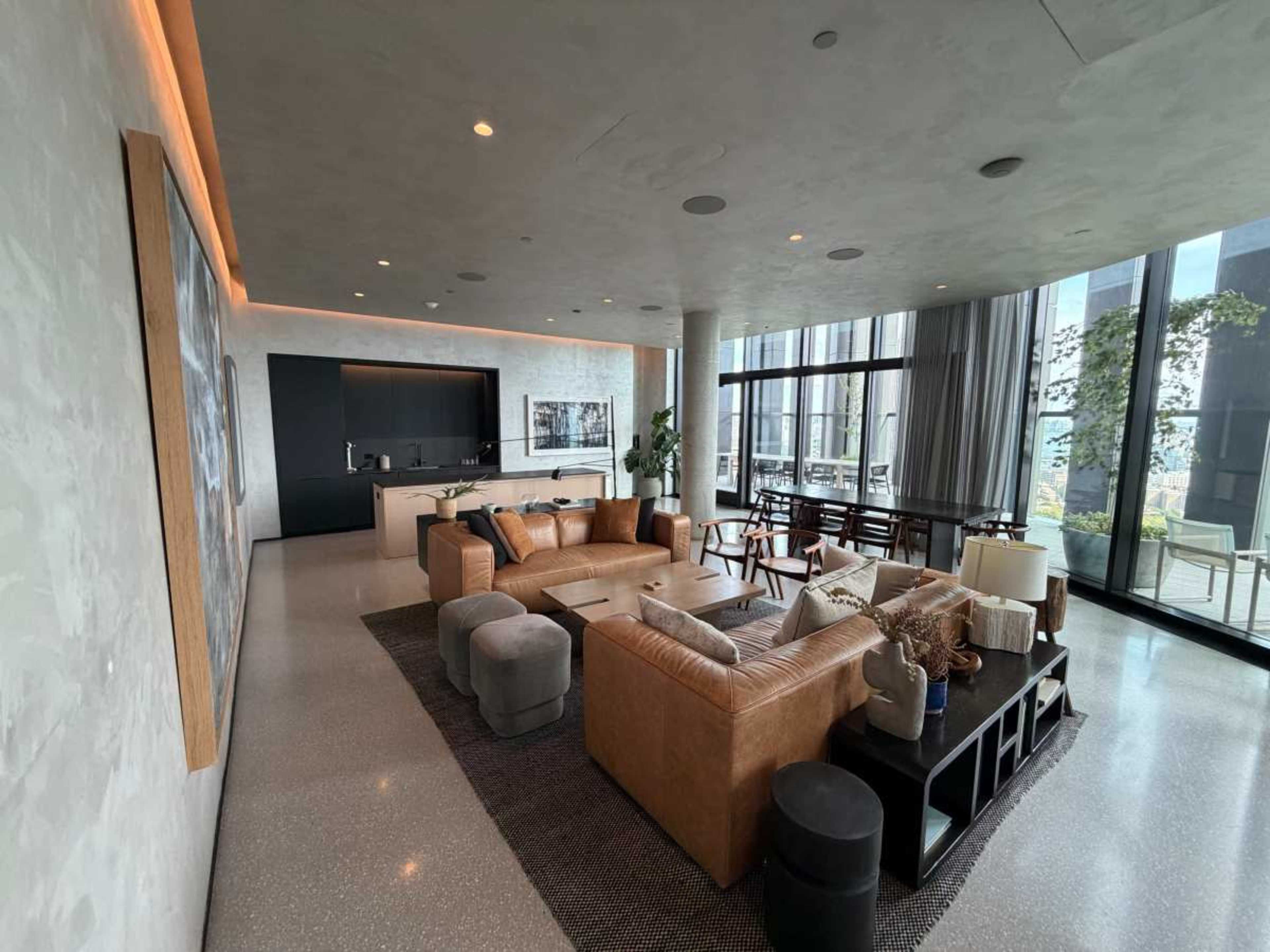 The image shows a spacious modern living room with large windows, a sectional leather couch, a dining area, and minimalist decor.