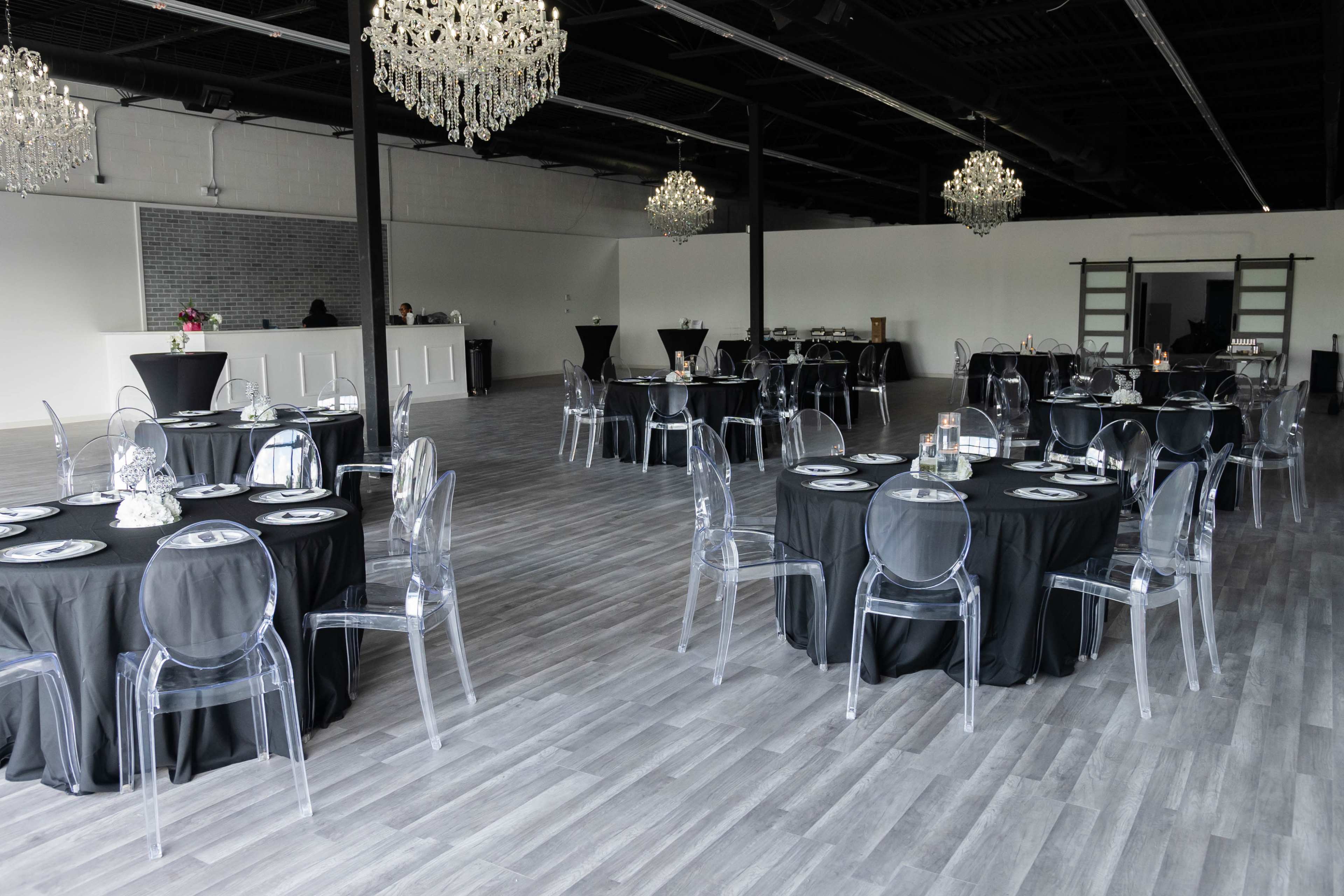 The image shows an elegantly arranged banquet hall featuring black tablecloths and clear acrylic chairs, with chandeliers hanging from the ceiling.