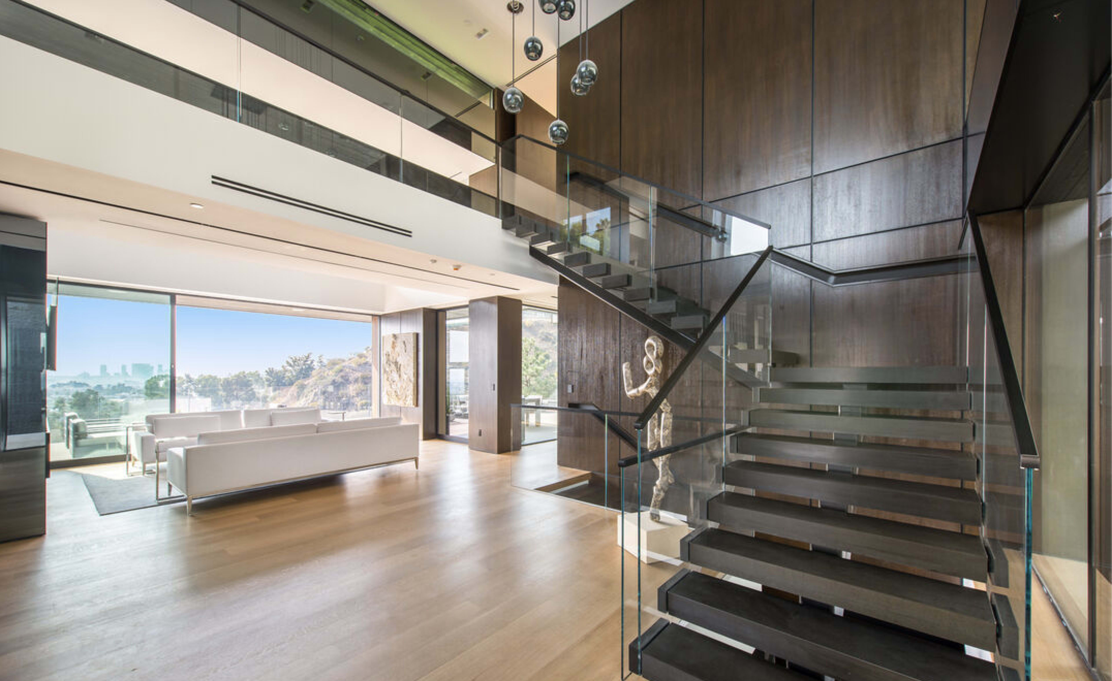 A modern interior features a wooden staircase with glass railings leading to an open living area that offers a view of the city skyline through large windows.