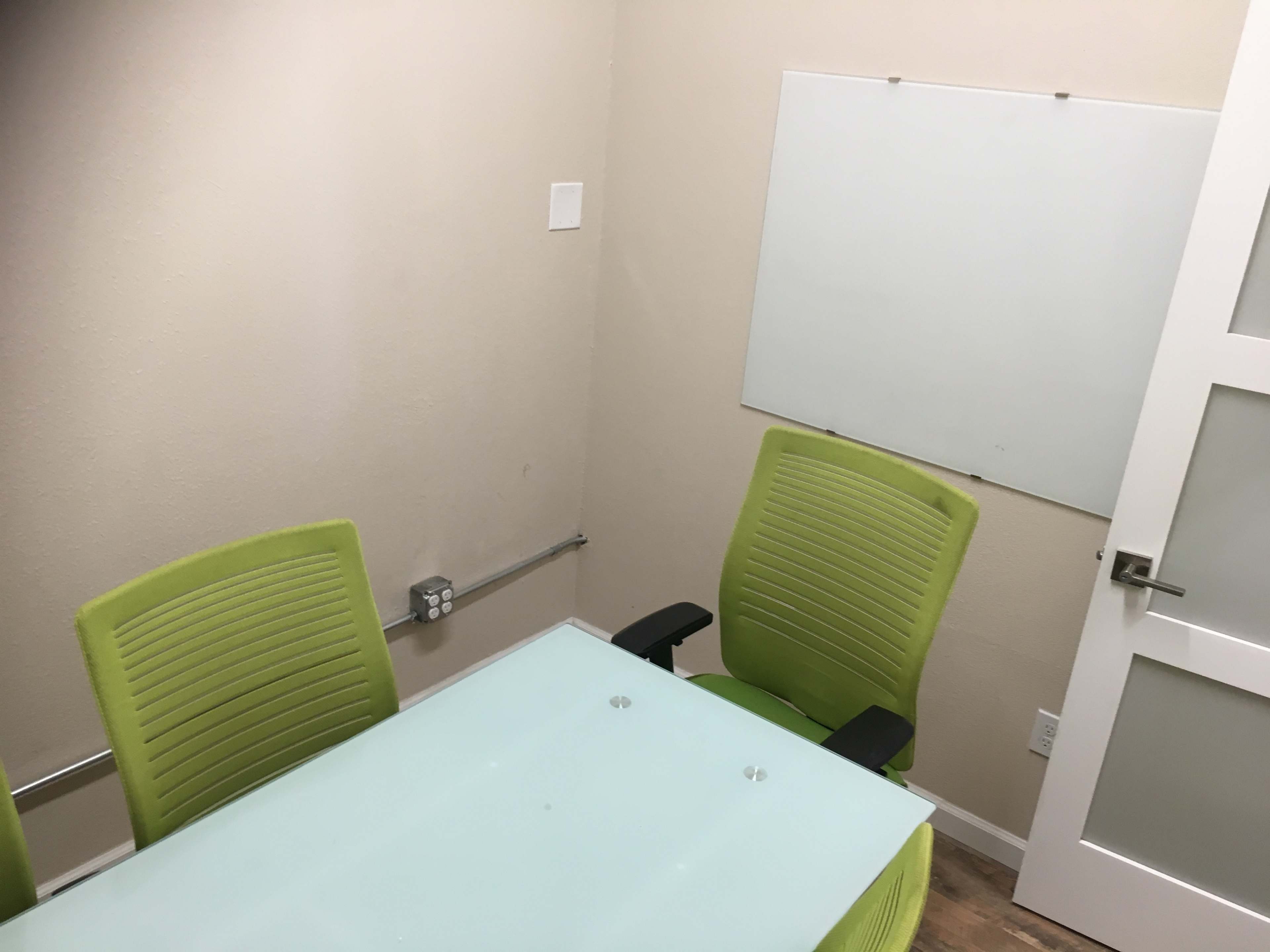 A small meeting room with a glass table, two green chairs, and a whiteboard attached to the wall.