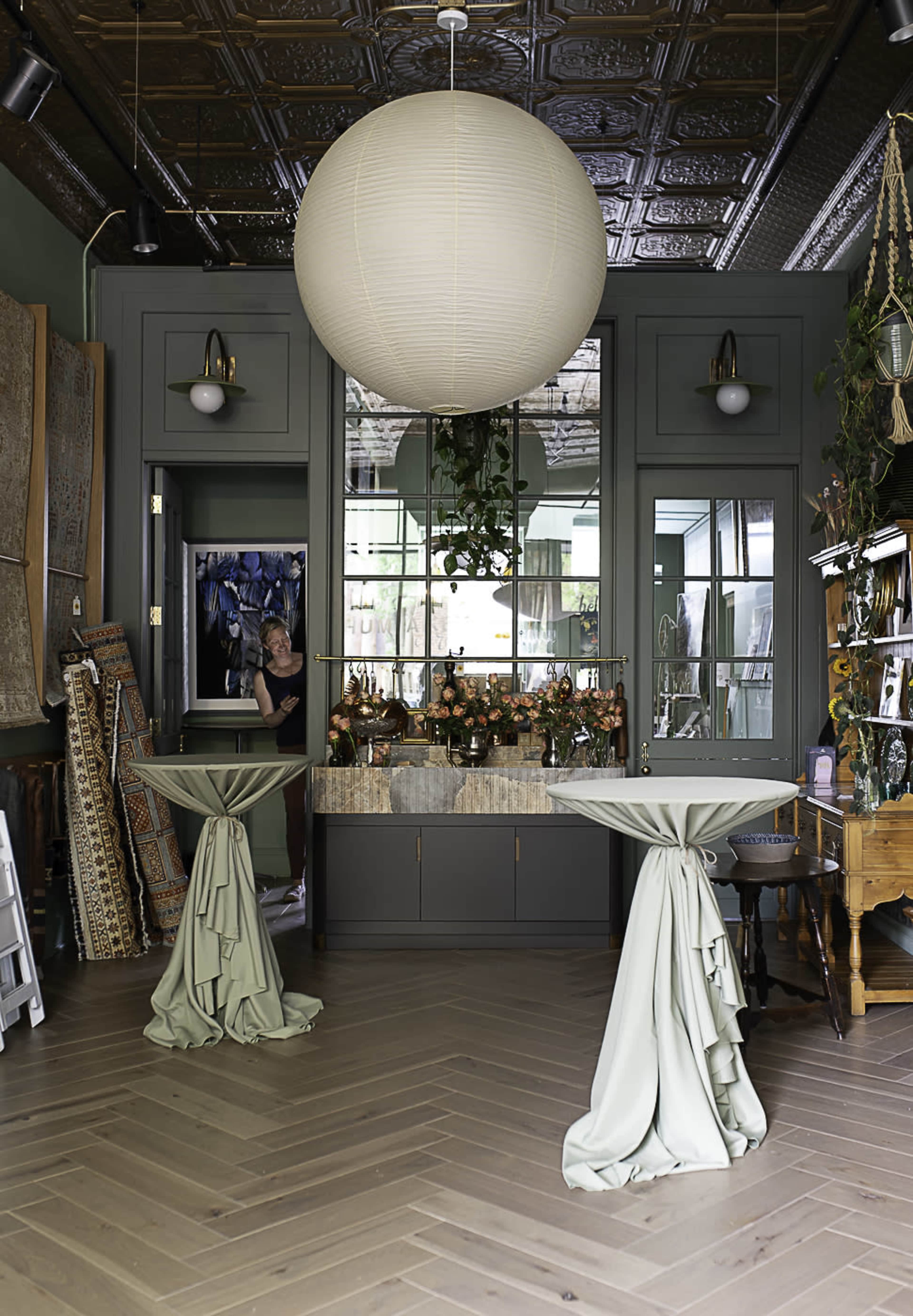 The image features a warmly styled interior with two draped tables, a large round light fixture, and a backdrop of greenery and decorative accents.