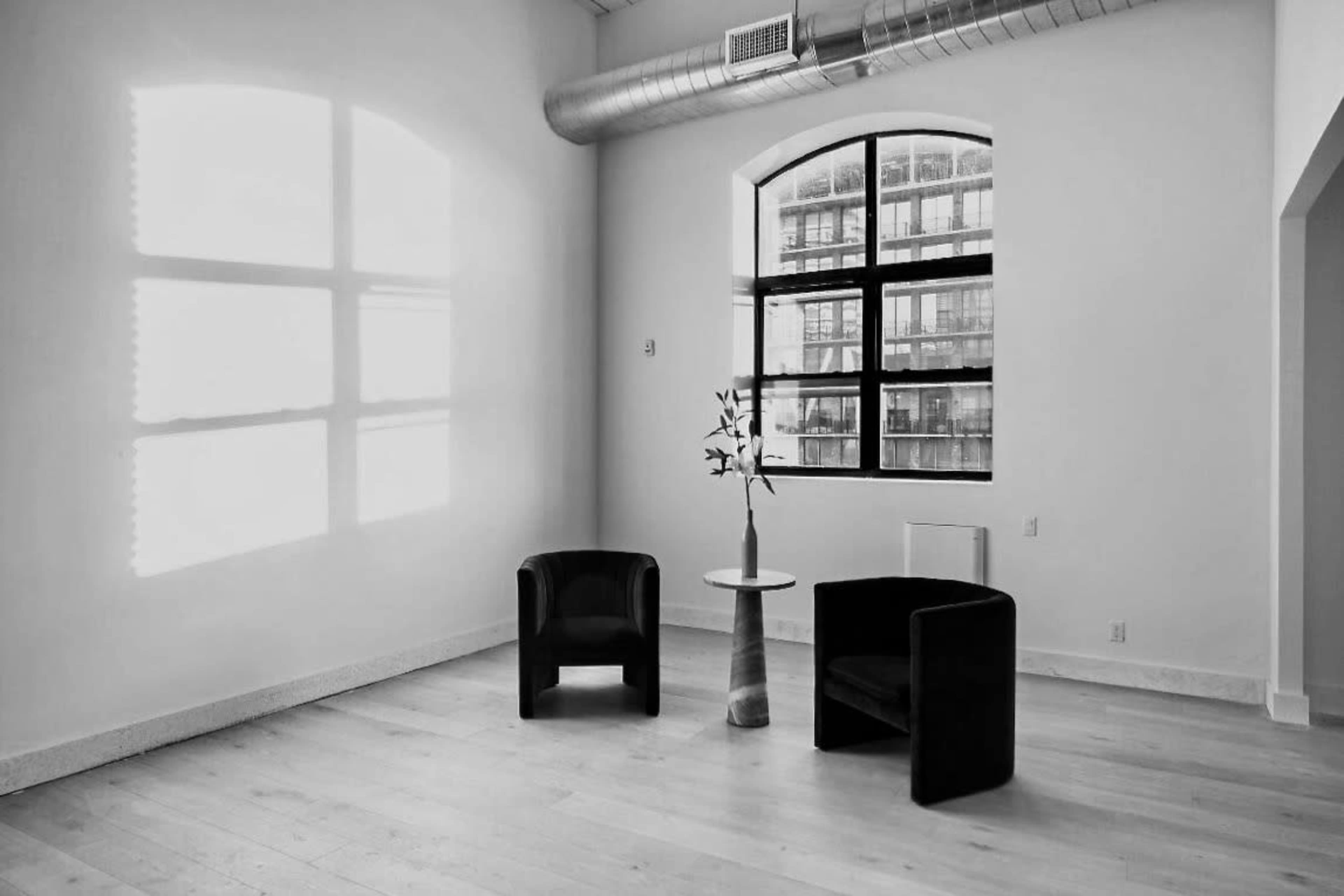 Two black chairs are positioned on a light wooden floor beside a small table with a vase, illuminated by a window casting shadows on the wall.