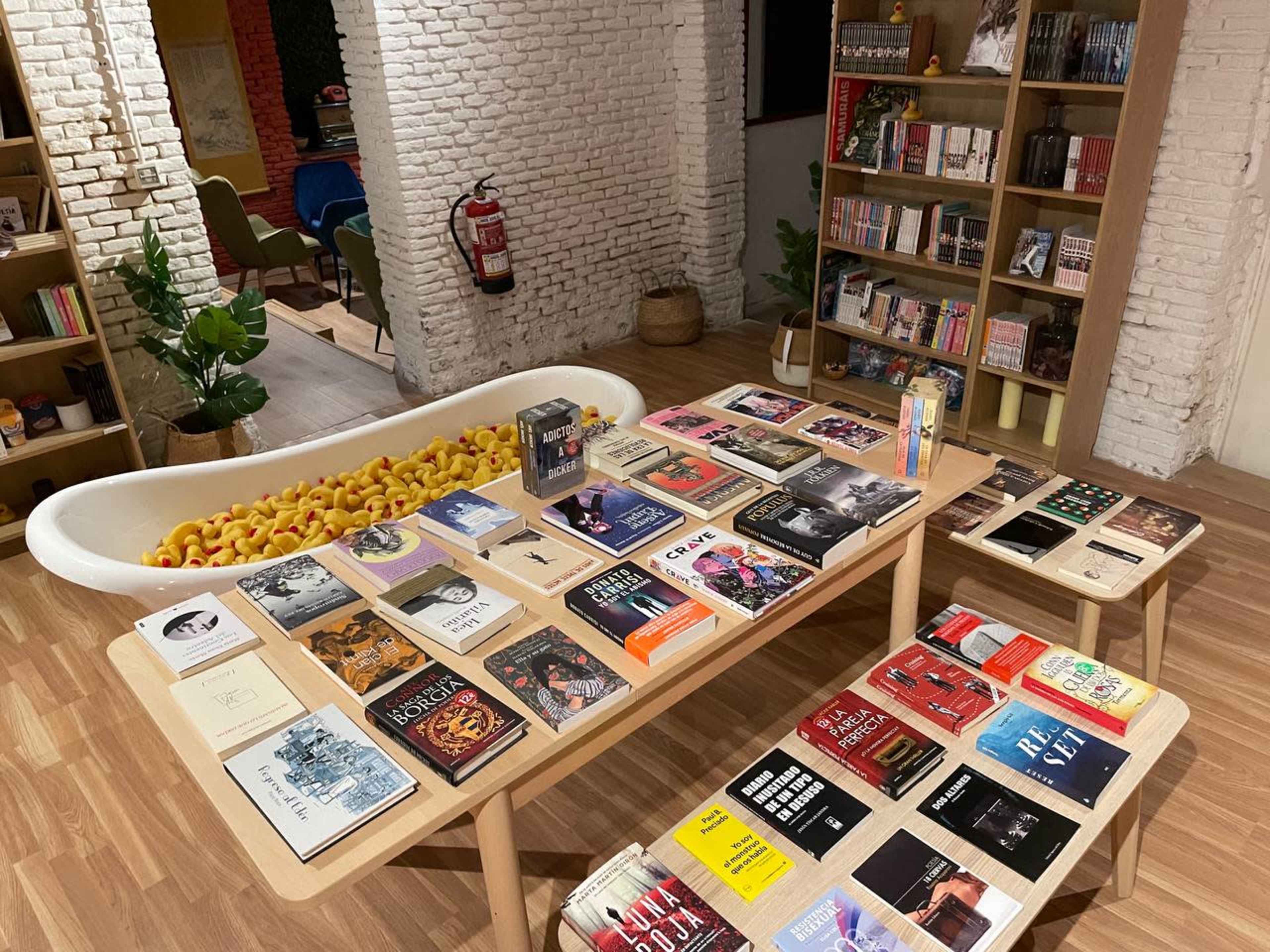 A cozy reading area features a wooden table filled with various books and a bathtub filled with rubber ducks in the background.