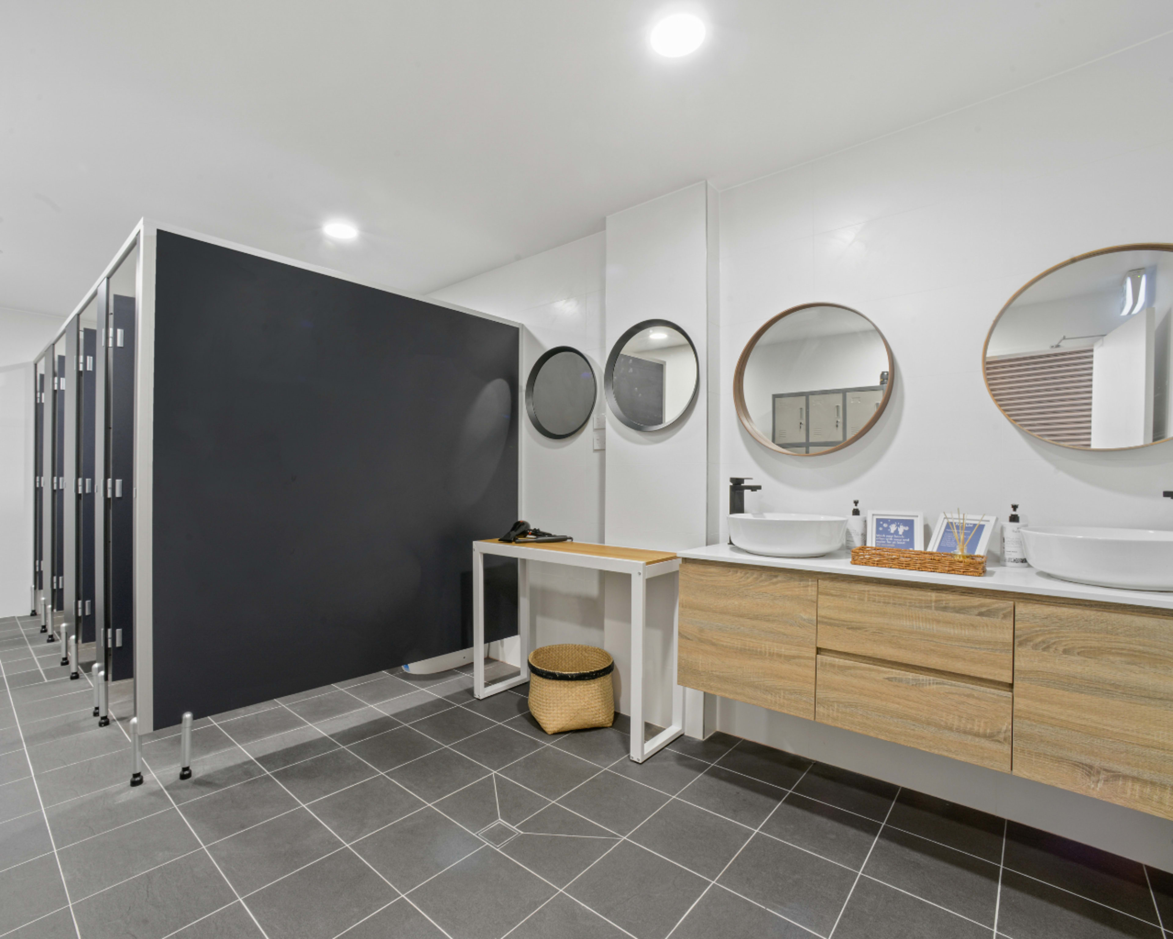The image shows a modern bathroom with private stalls, round mirrors, and a wooden vanity with two sinks.