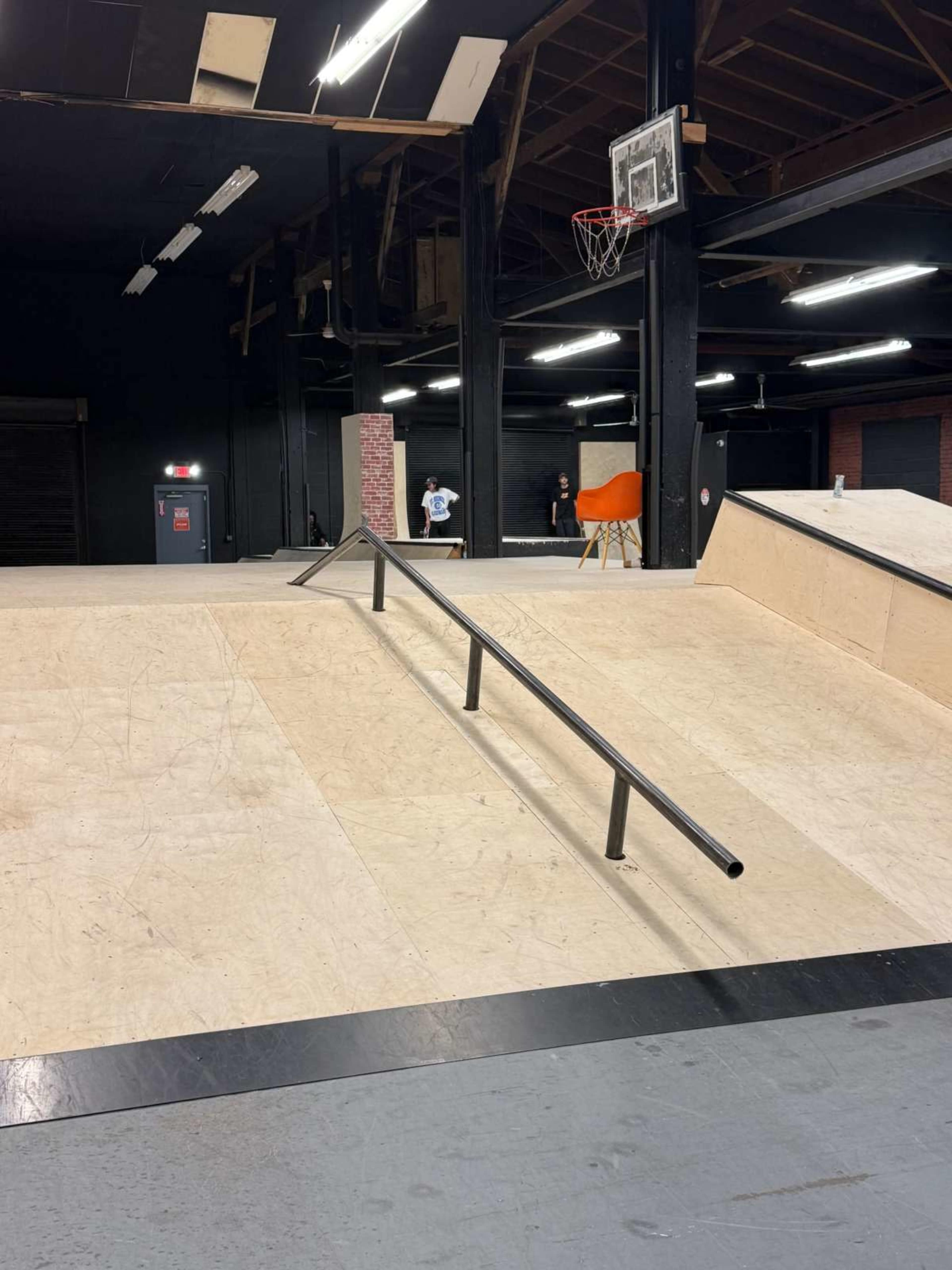 The image shows a skatepark interior featuring a wooden ramp with a metal railing and a basketball hoop in the background.
