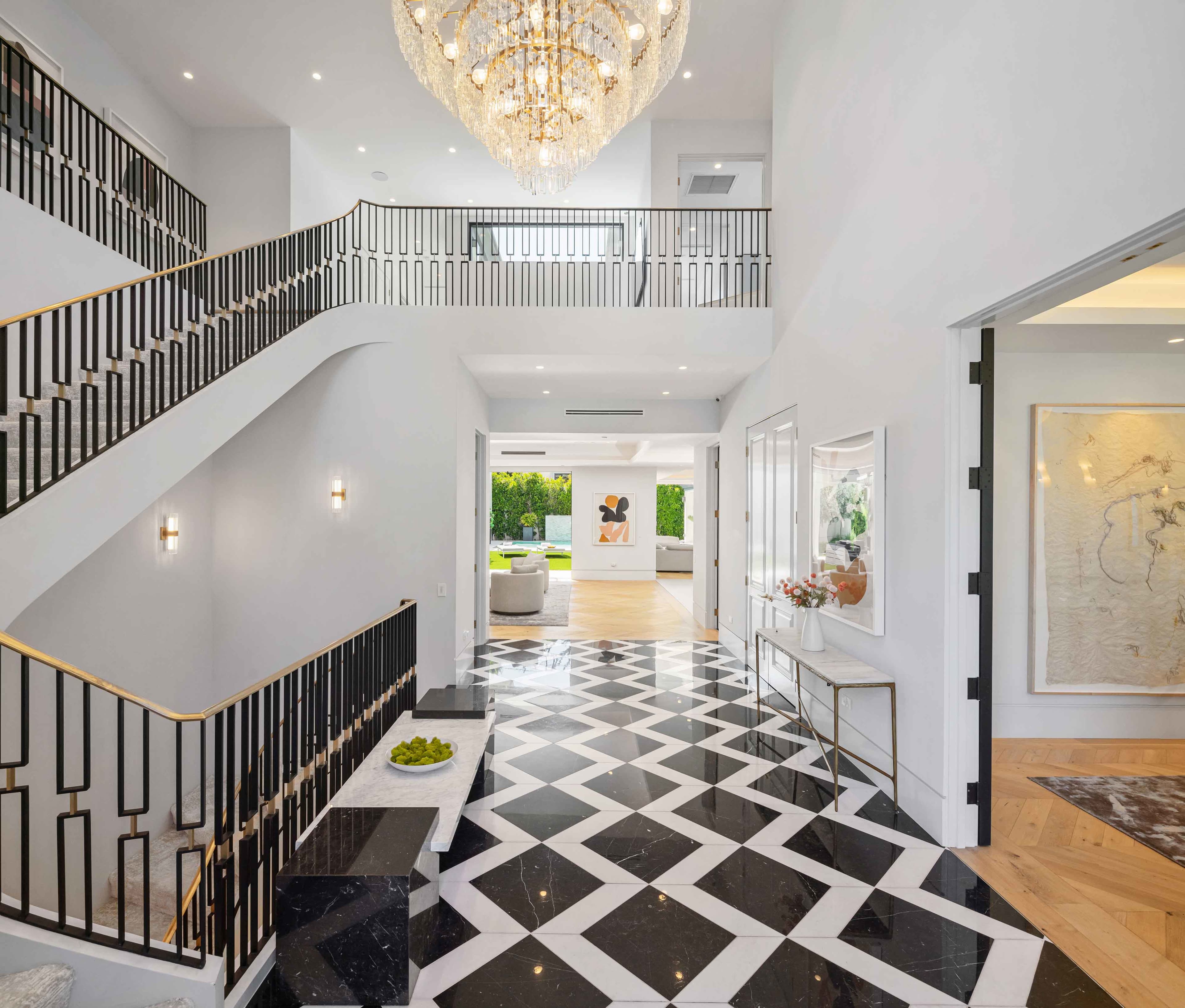 The image shows a modern foyer with a grand staircase, featuring a geometric black-and-white tiled floor and a large chandelier overhead.