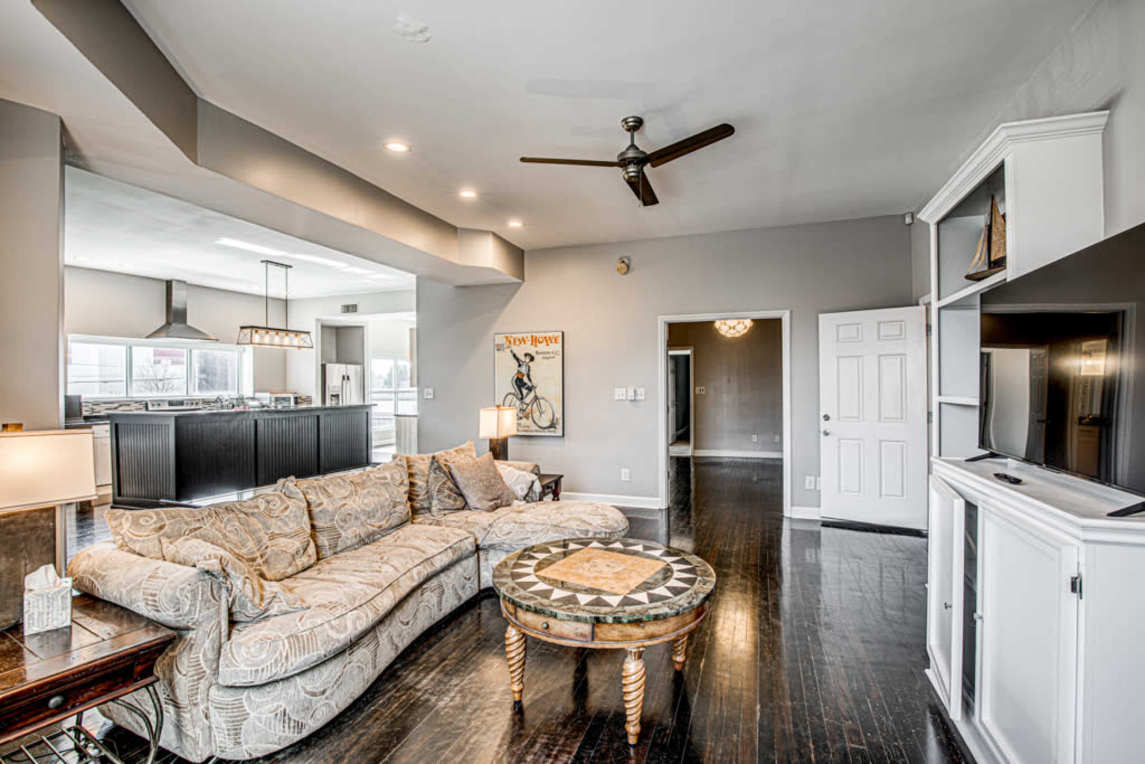 The living room features a large sectional sofa, a decorative coffee table, and an open layout leading to a kitchen area in the background.