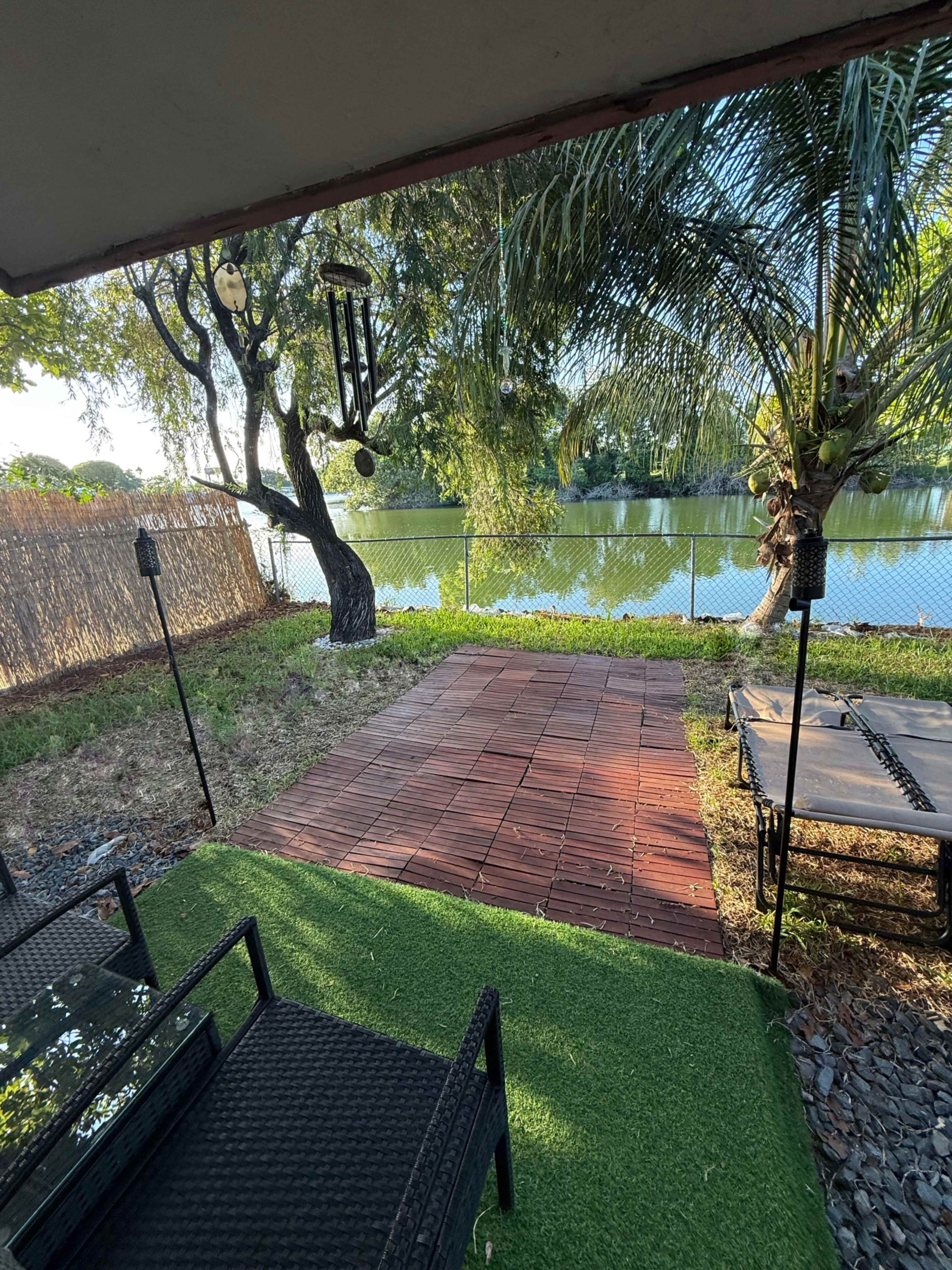 The image shows a patio area overlooking a calm body of water, with a wooden deck, green grass, and palm trees.