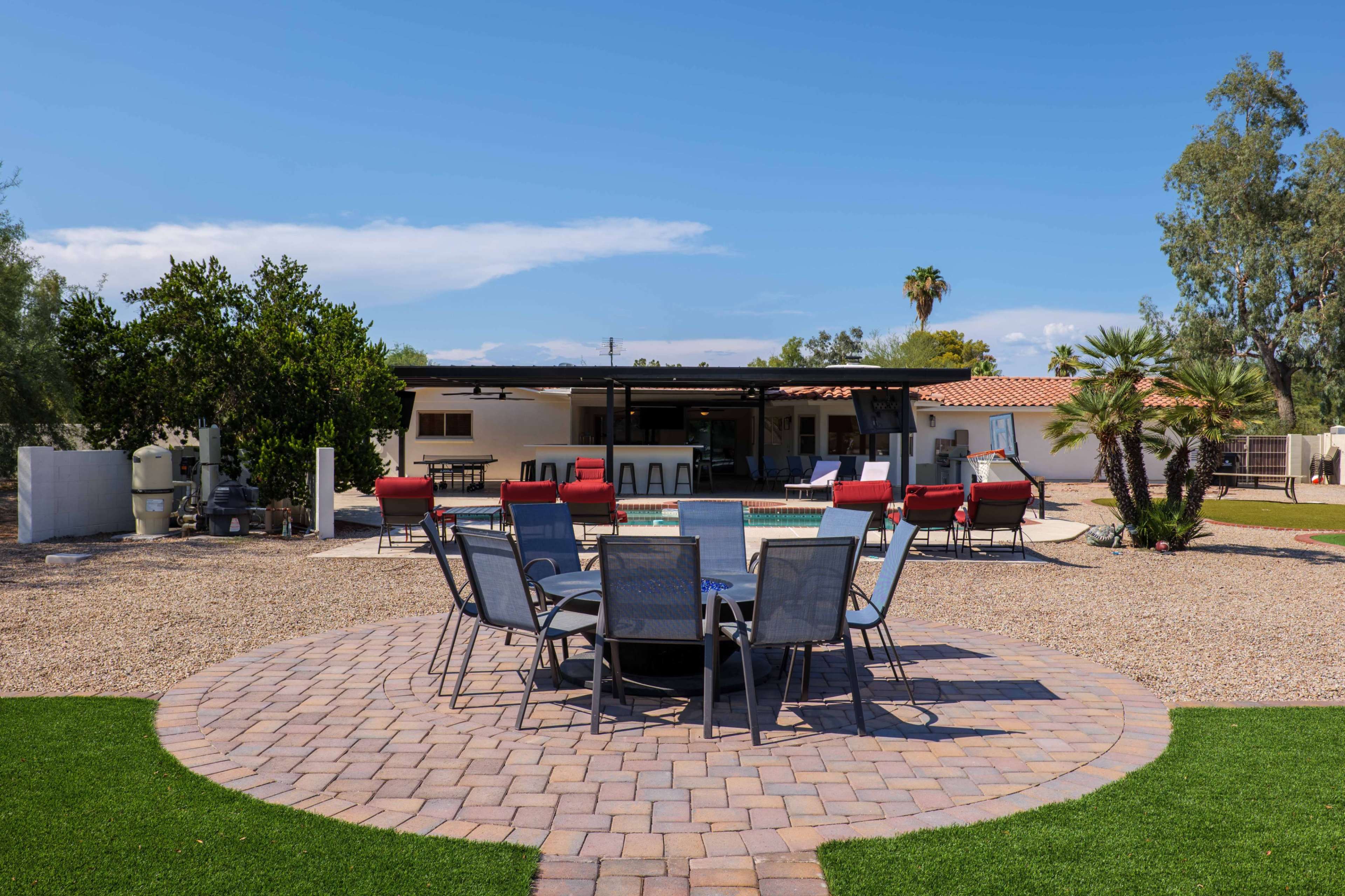 The image shows a landscaped backyard with a circular stone patio surrounded by chairs, a pool, and a house in the background.