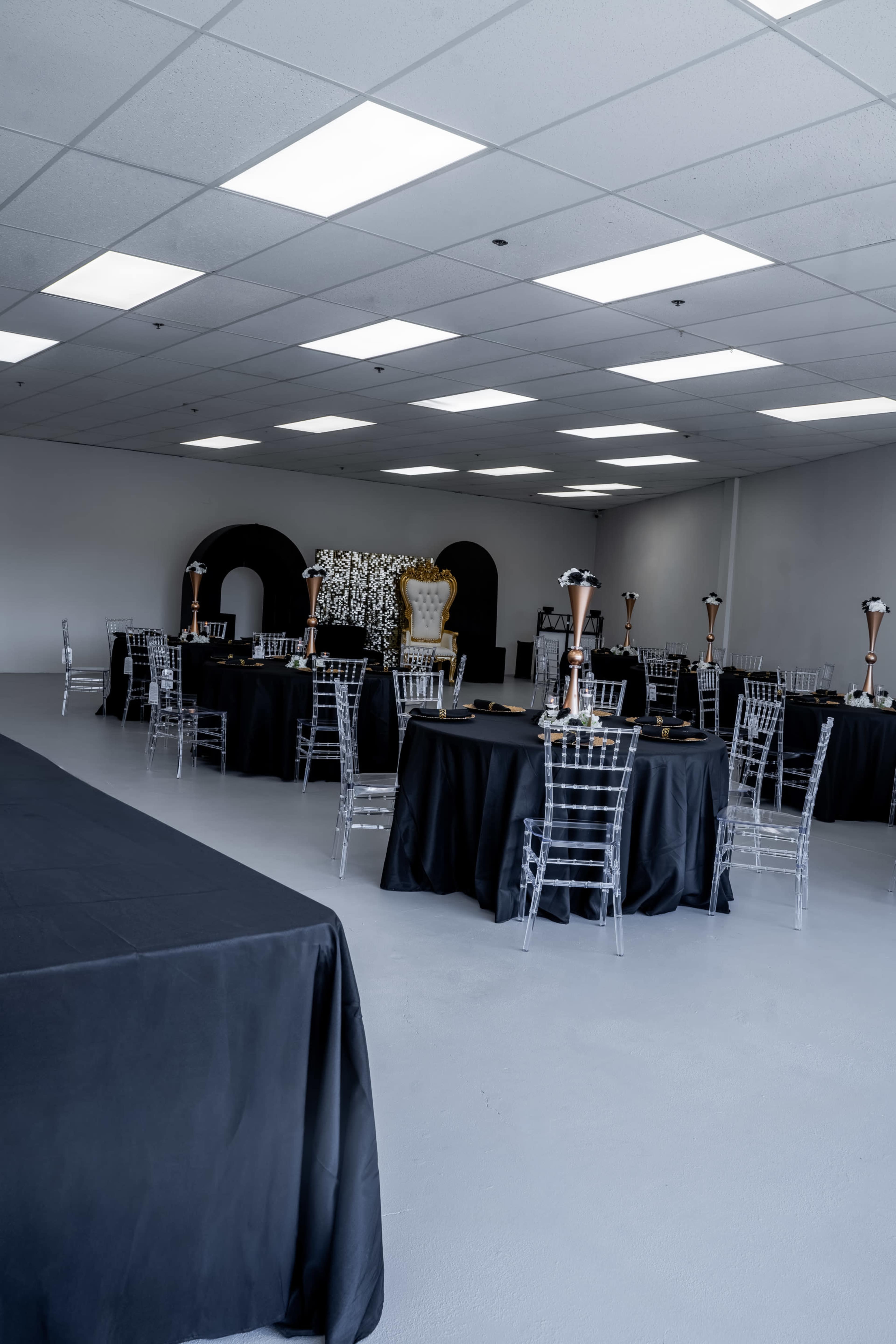 An empty event space features several round tables dressed in black tablecloths, each adorned with clear chairs and elegant centerpieces.
