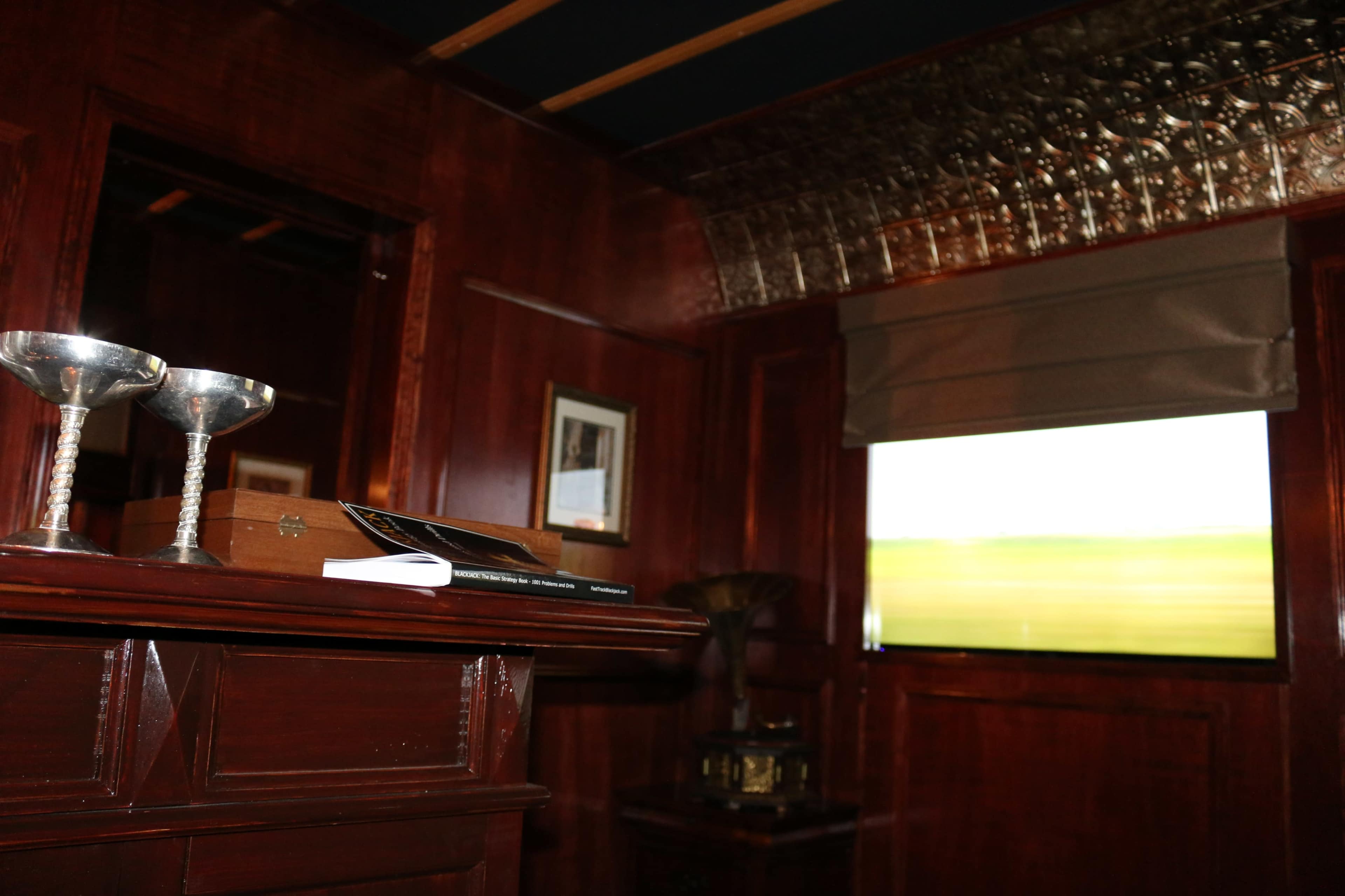 The image depicts a wooden interior with a bar area featuring two silver chalices and a window displaying a blurred green landscape.