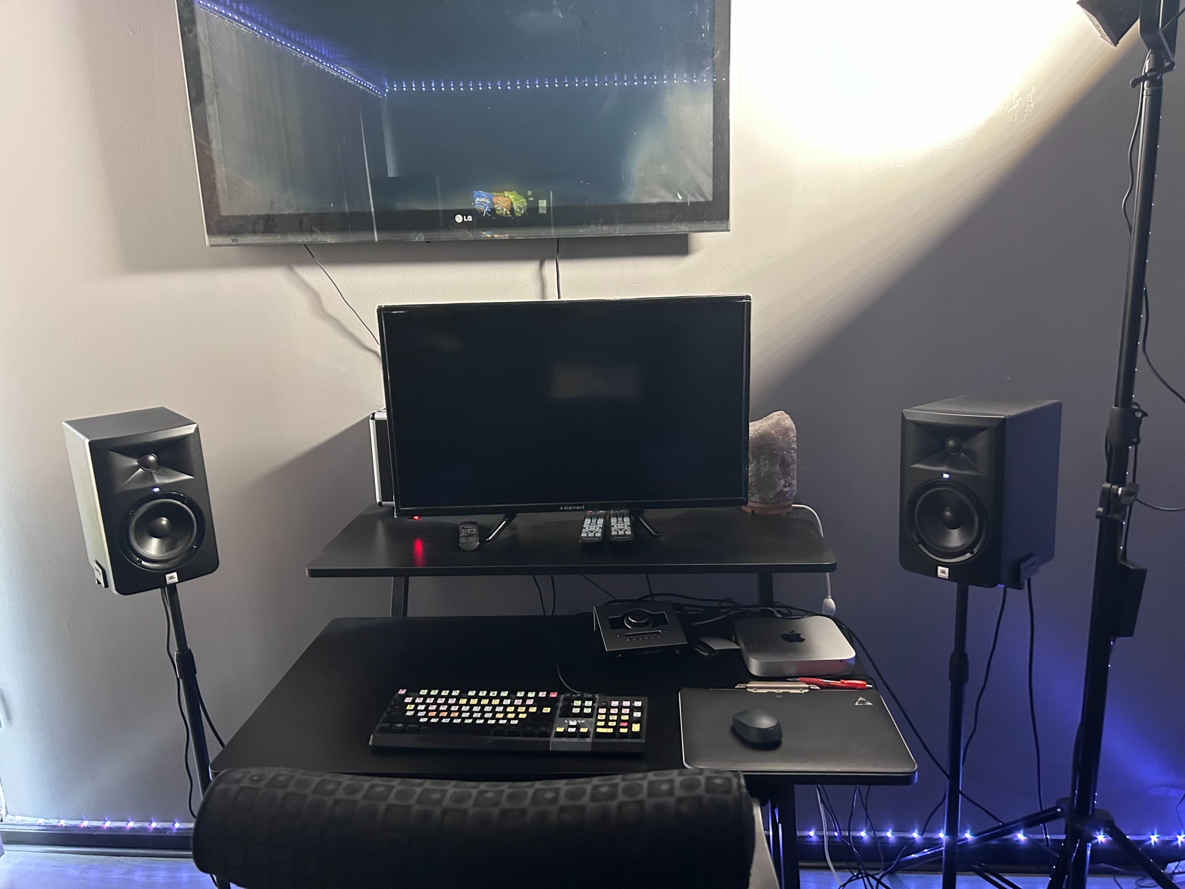 A black desk with a computer monitor, keyboard, remote, and speakers is positioned against a wall, illuminated by soft lights.