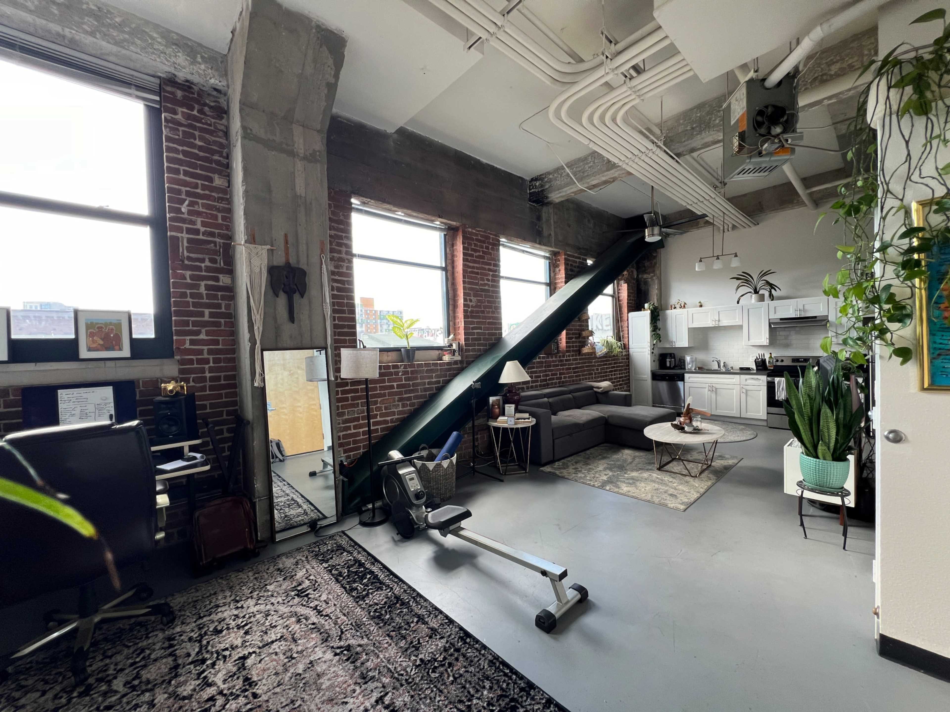 The image shows a spacious loft-style apartment featuring large windows, exposed brick walls, a seating area with a gray sofa, a kitchen area, and workout equipment.