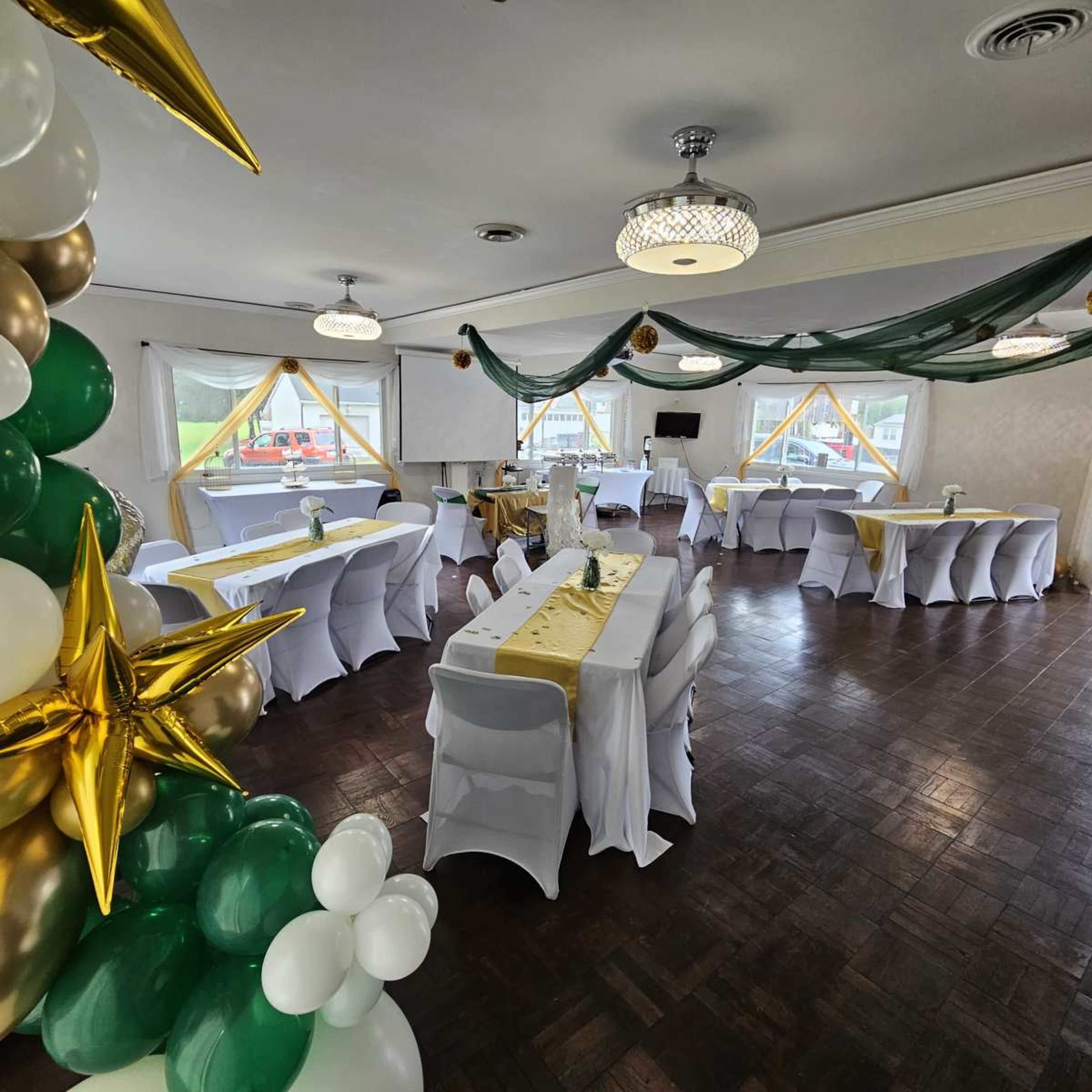 The image shows an event space set up with tables covered in white tablecloths, decorated with gold and green accents, and adorned with balloons and draped fabric.