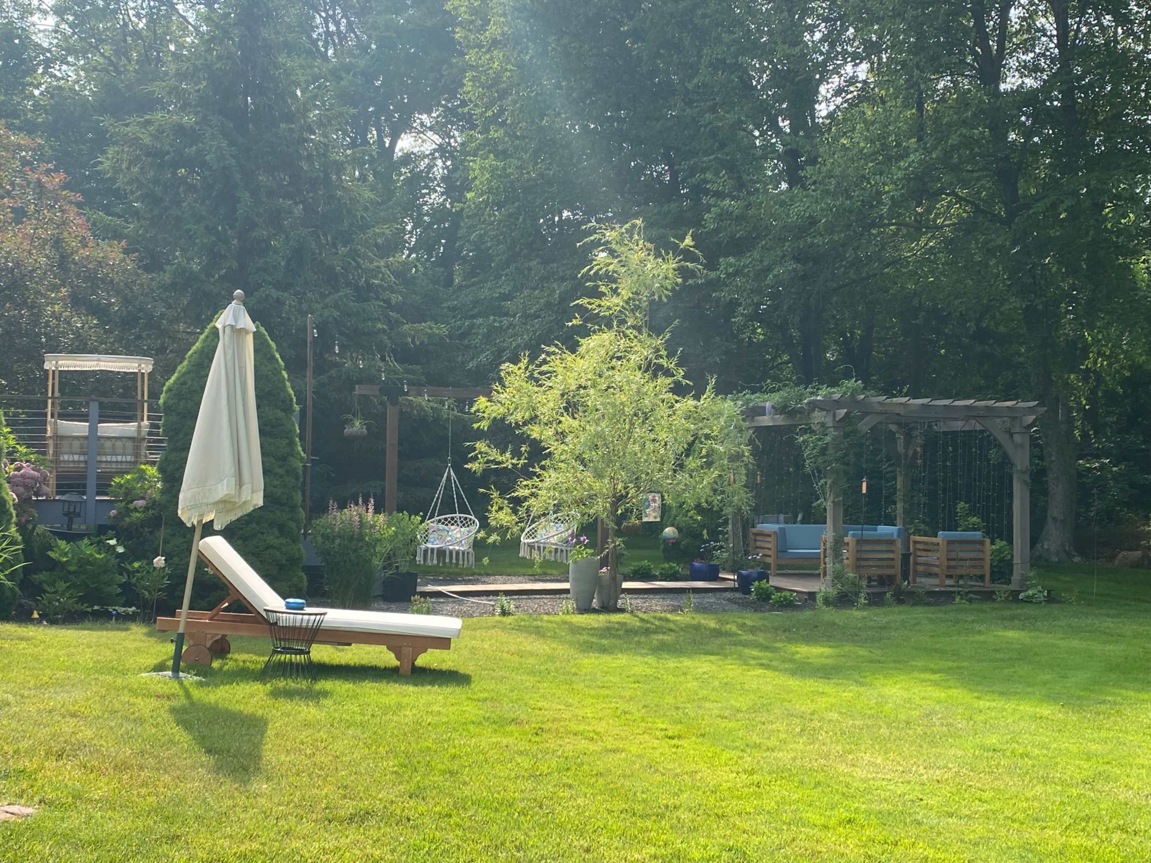 The scene shows a well-kept garden with a lounge chair, an umbrella, swings, and a pergola surrounded by greenery.