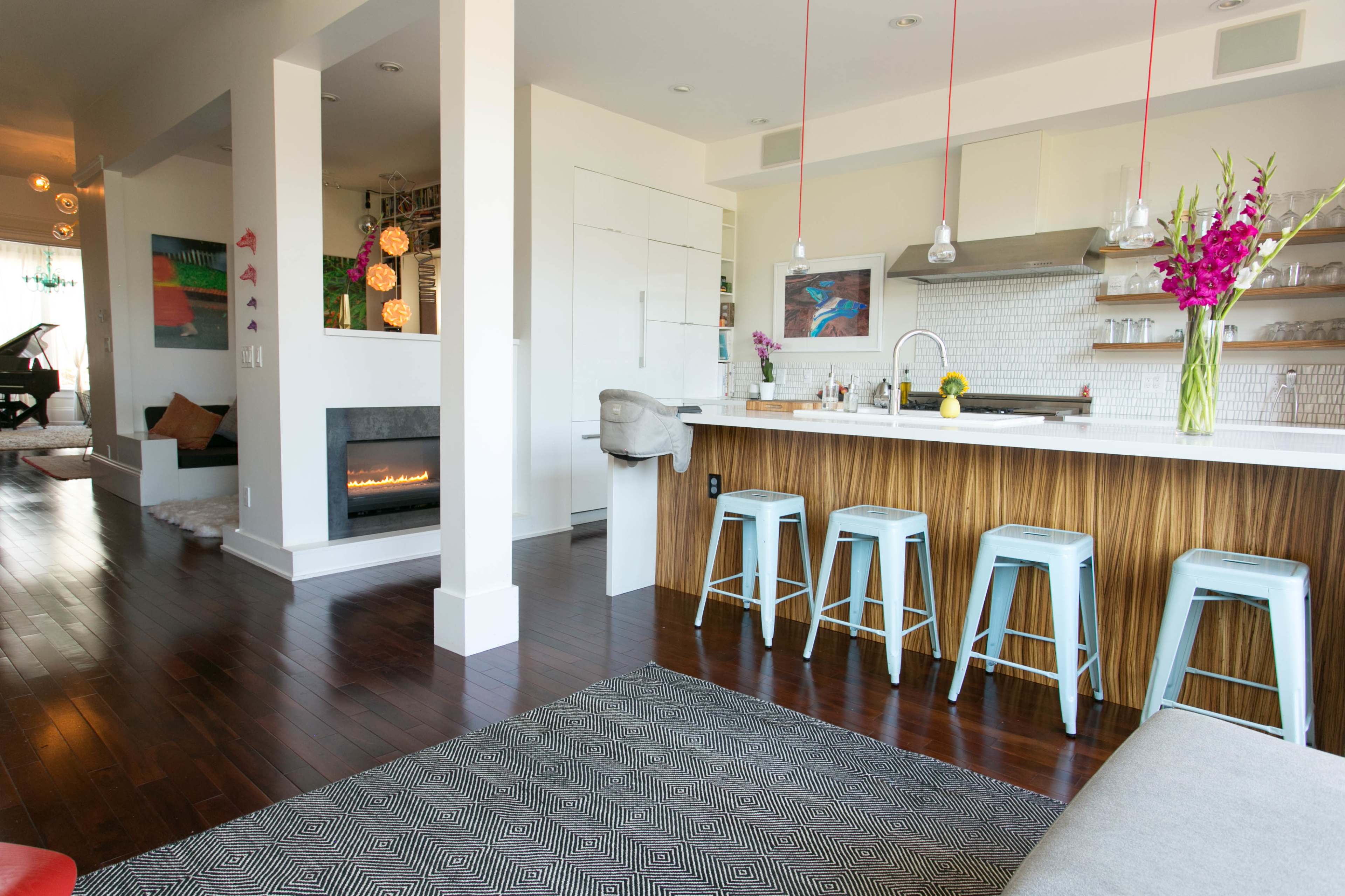 A modern open-concept living space featuring a kitchen with a bar, stools, and a fireplace, alongside a living area decorated with art and ambient lighting.