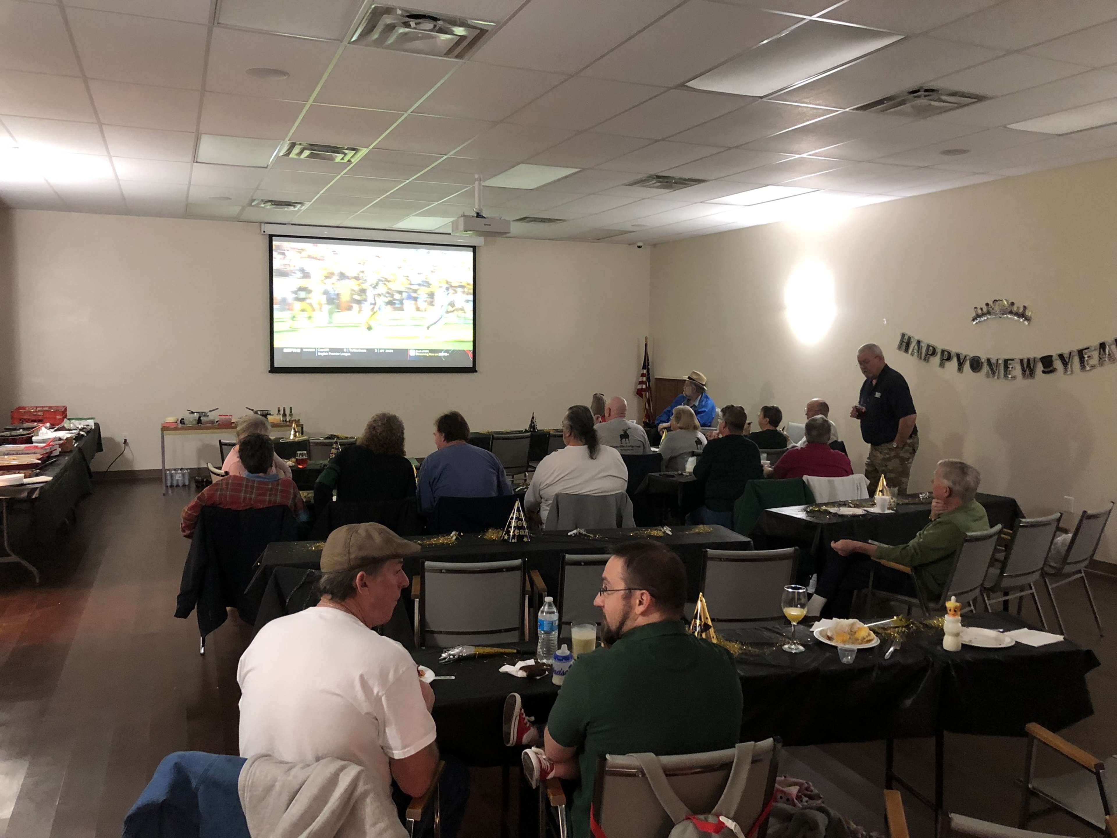 A group of people watches a sports event on a large screen in a decorated room with tables set for a gathering.