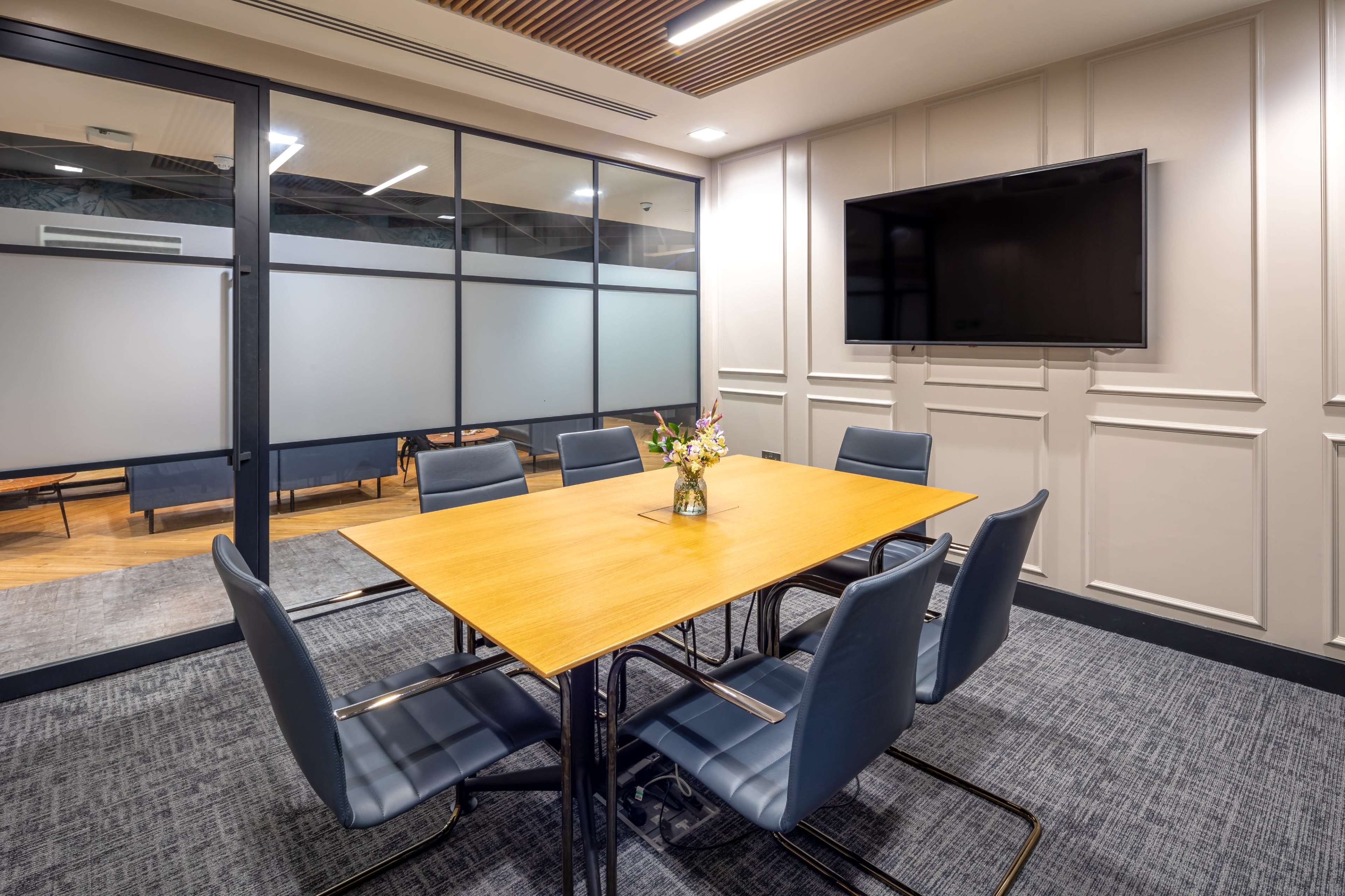 A modern conference room features a large wooden table surrounded by six blue chairs, with a wall-mounted television and a flower vase at the center.