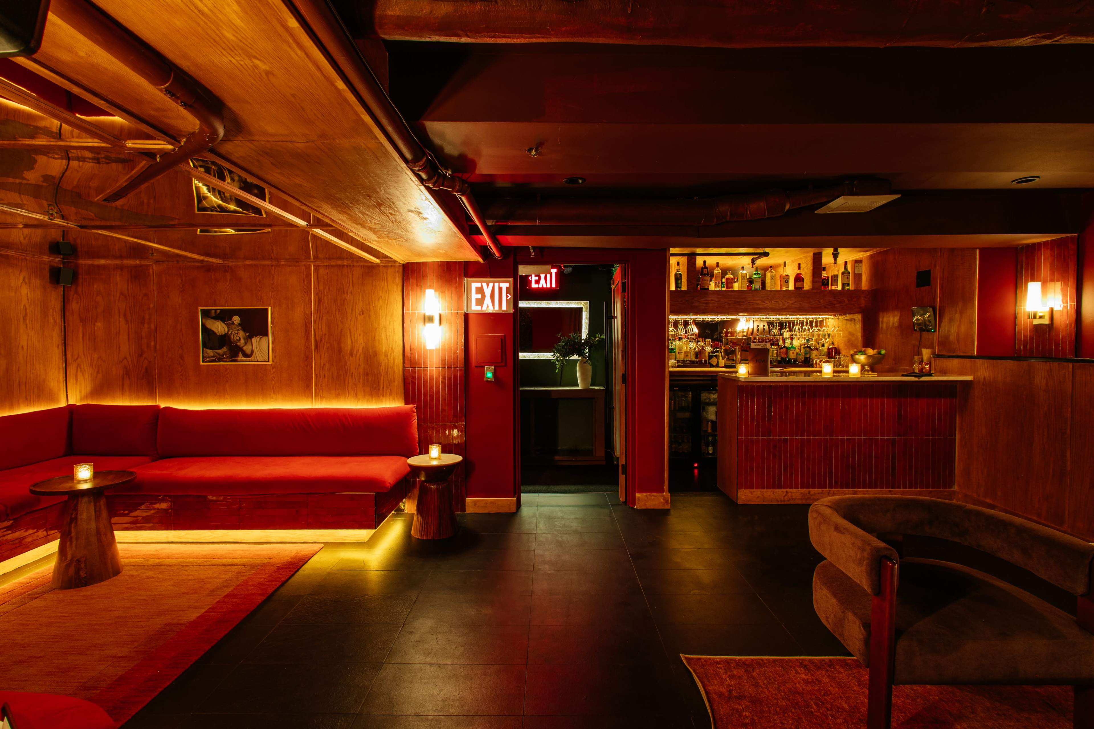 The image shows a dimly lit bar interior featuring red seating, wooden paneling, and a well-stocked bar area in the background.