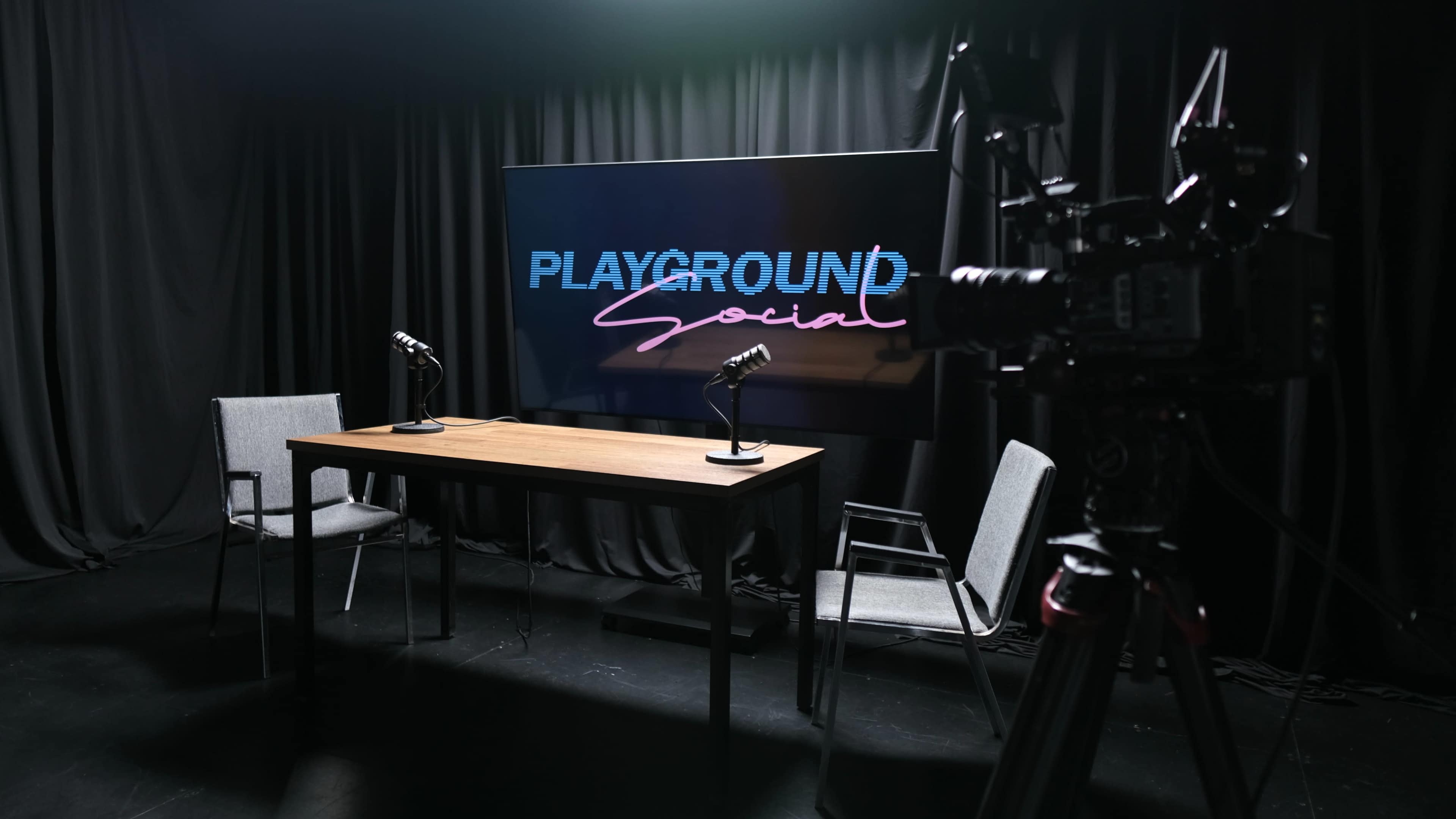 A modern studio setup features a wooden table with two chairs, microphones, and a large screen displaying the text "PLAYGROUND Social."